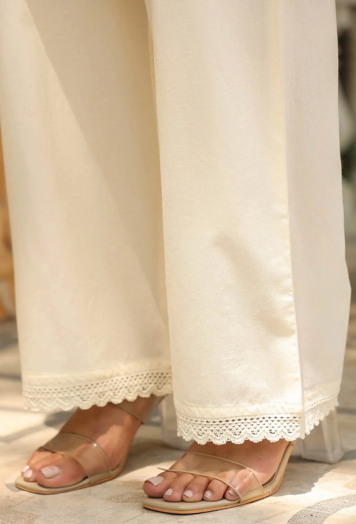 Close-up of white pants with lace trim worn with beige sandals on a blurred background