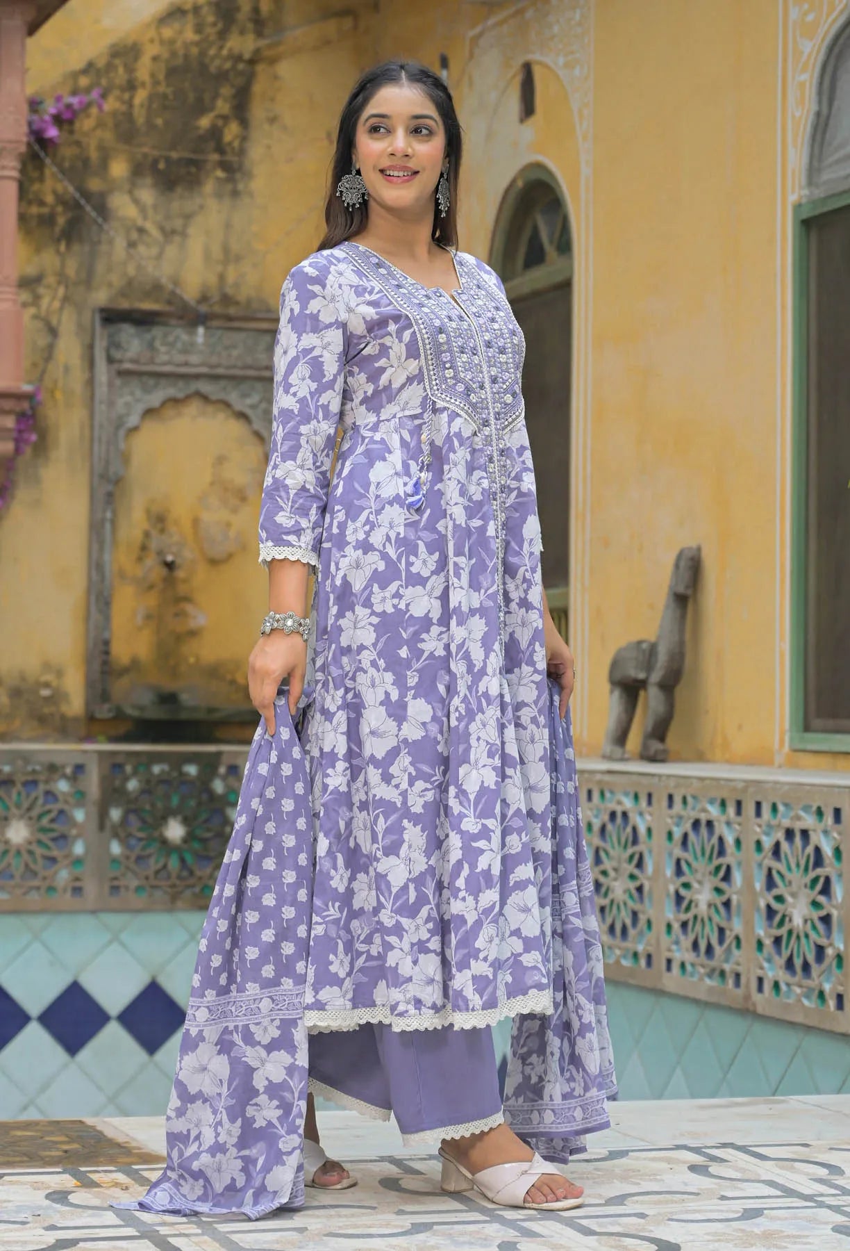 Woman wearing a purple floral outfit standing in front of a decorative wall.