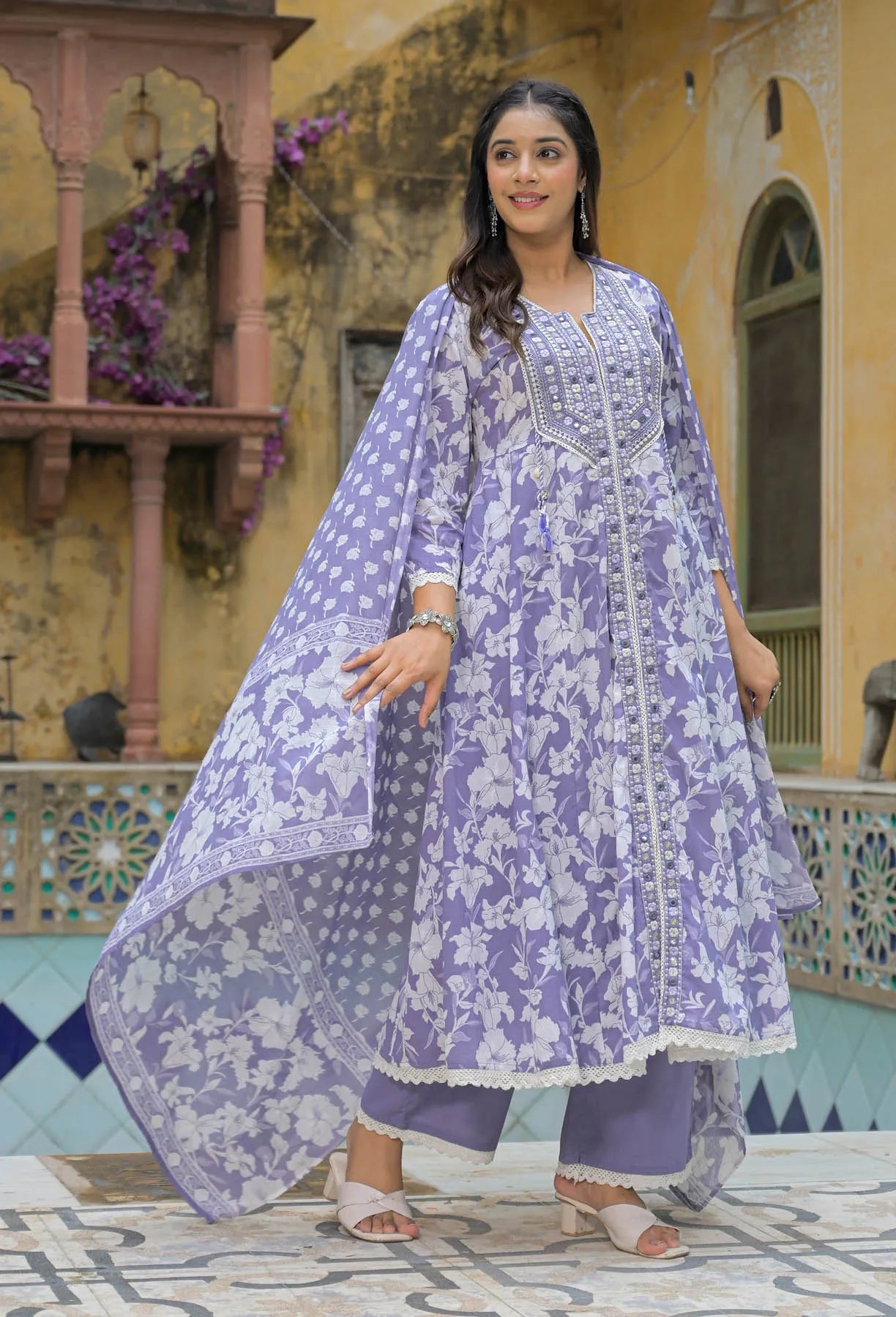 Woman wearing a purple floral traditional outfit in an outdoor setting with decorative architecture.