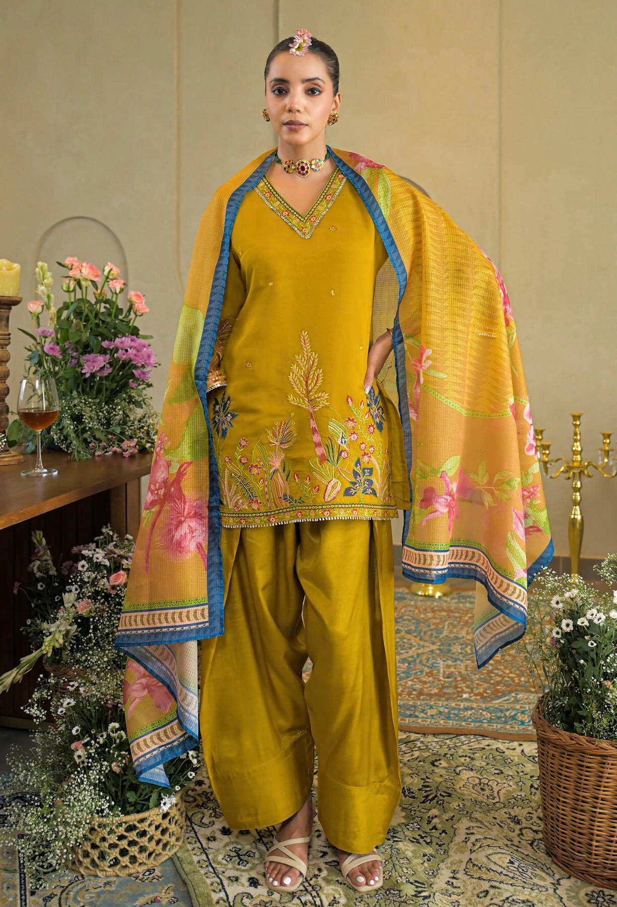 Woman in a mustard yellow traditional outfit with floral patterns in an elegant indoor setting.