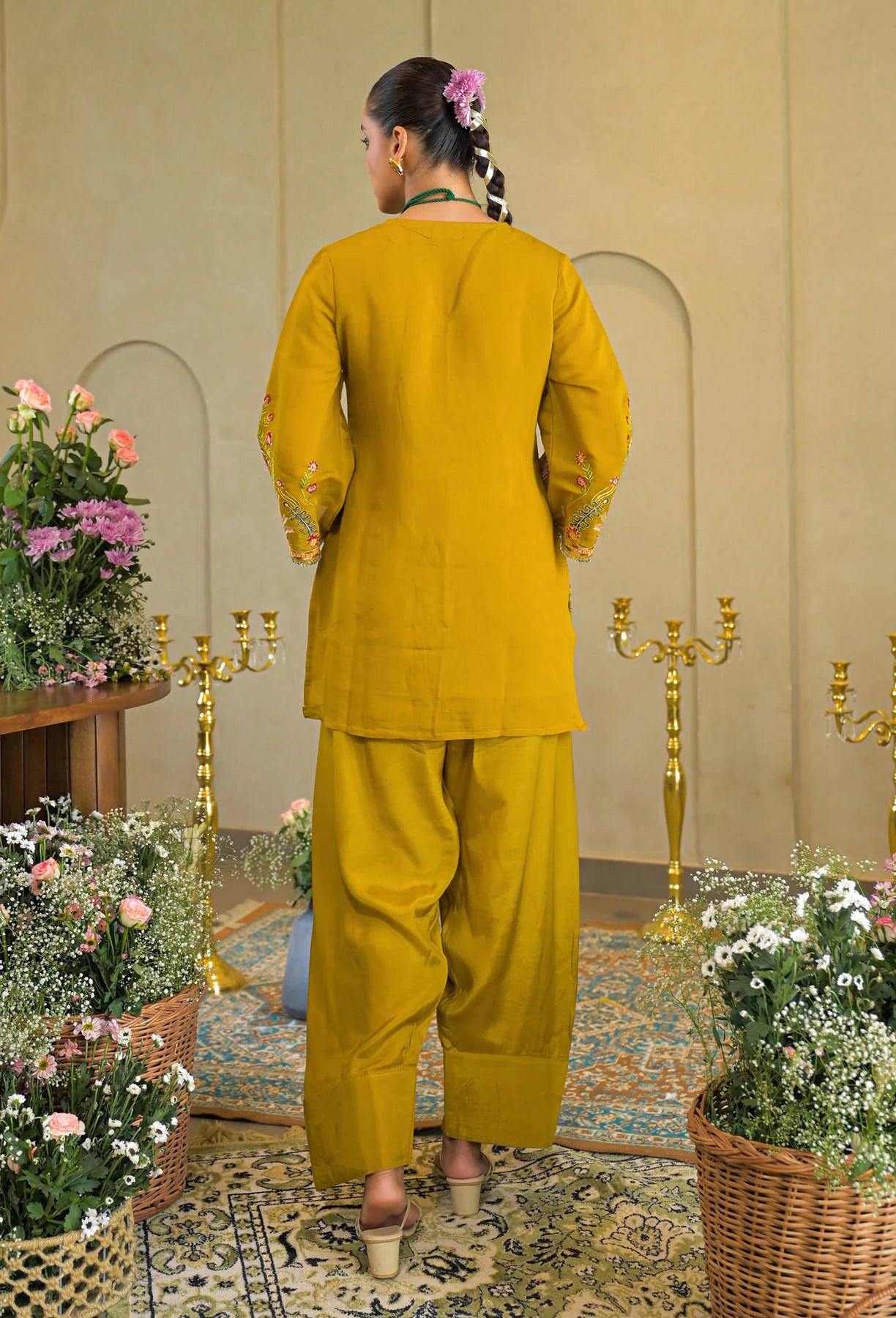 Woman in a mustard yellow traditional outfit standing in a decorated room with flowers and candles.