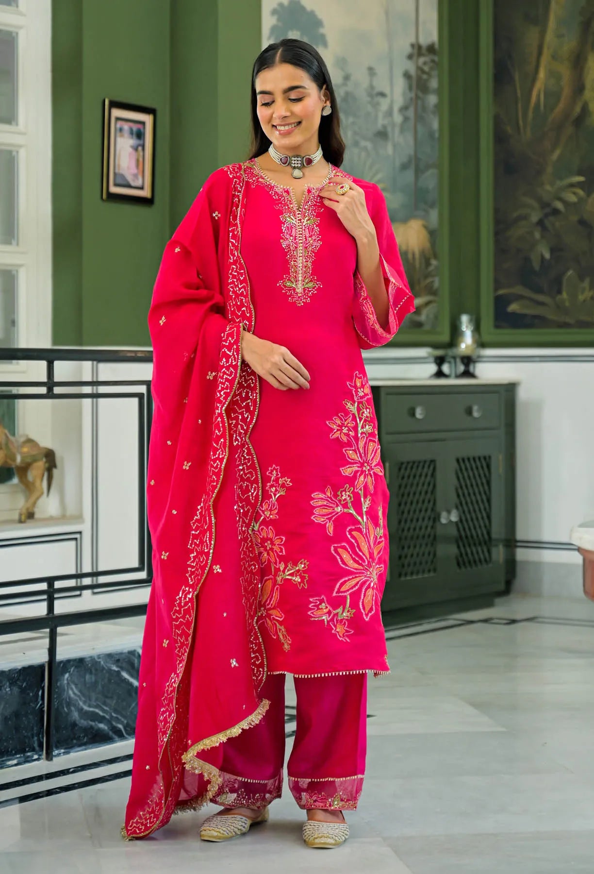Woman in a bright pink traditional outfit with embroidery in a room with green walls and decorative items.