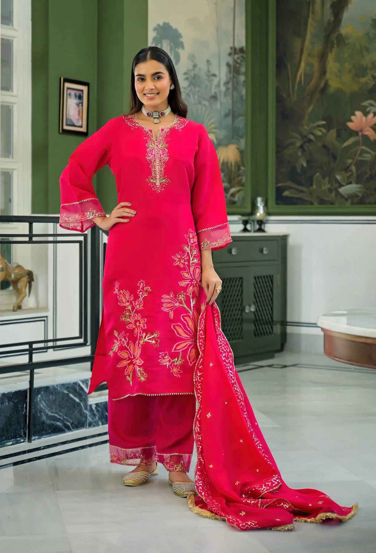 Woman in a bright pink traditional outfit with floral embroidery in a decorated room.