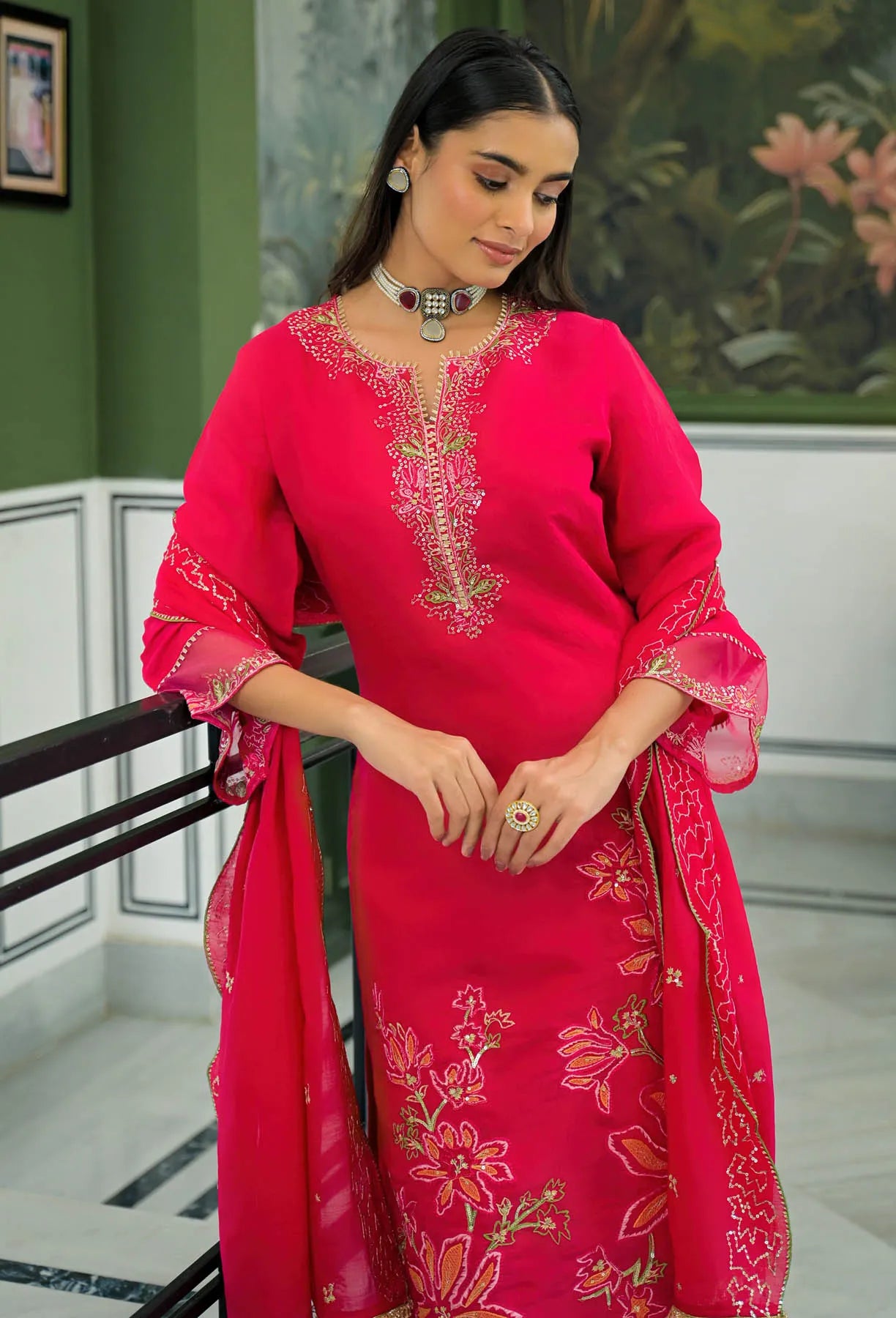 Woman wearing a bright pink traditional outfit with intricate designs in a decorated room.
