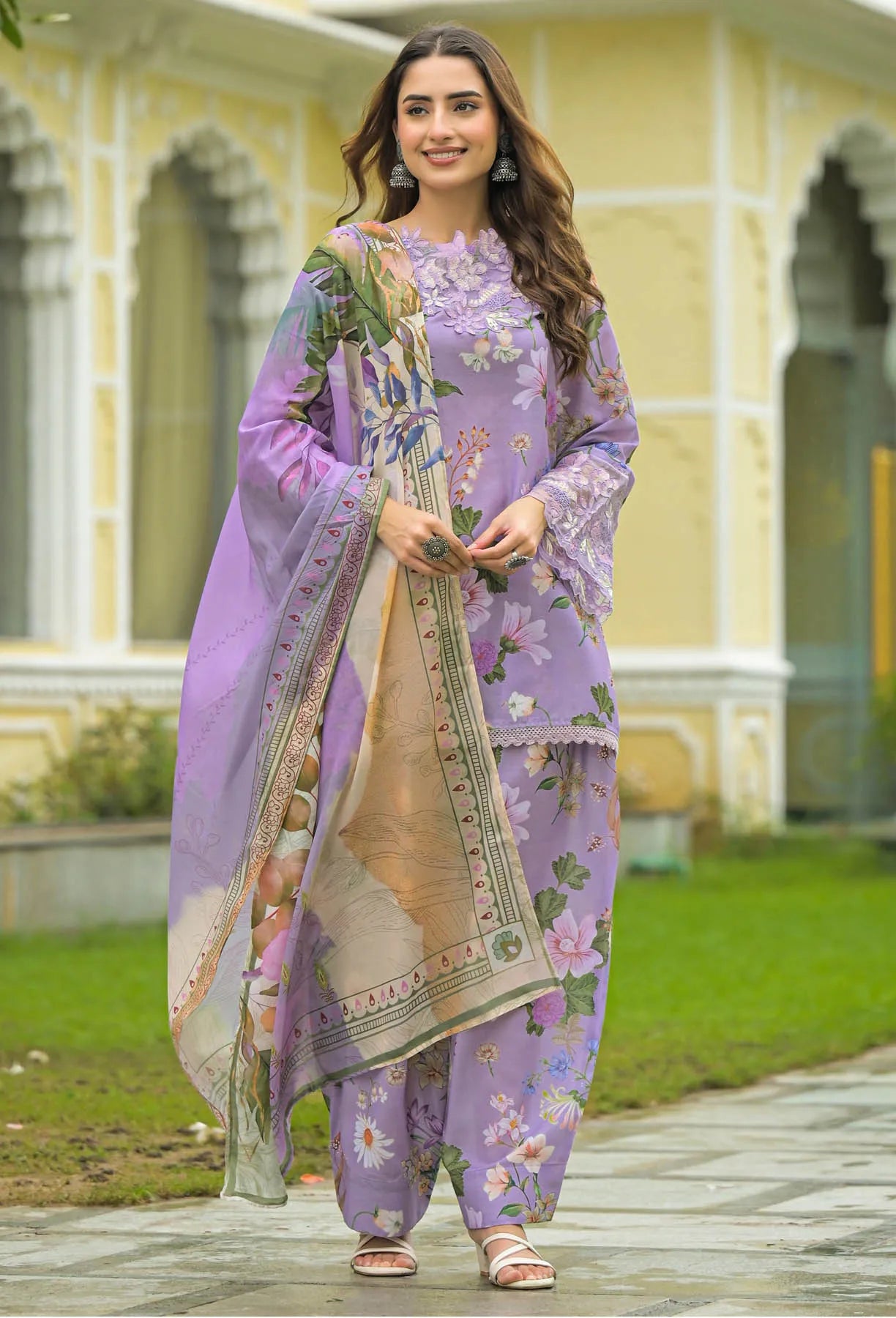 Woman in a purple floral outfit standing in front of a building with greenery.