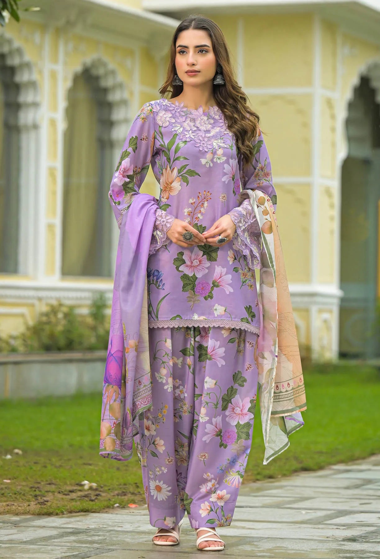 Woman wearing a floral purple outfit standing in front of a building with greenery.