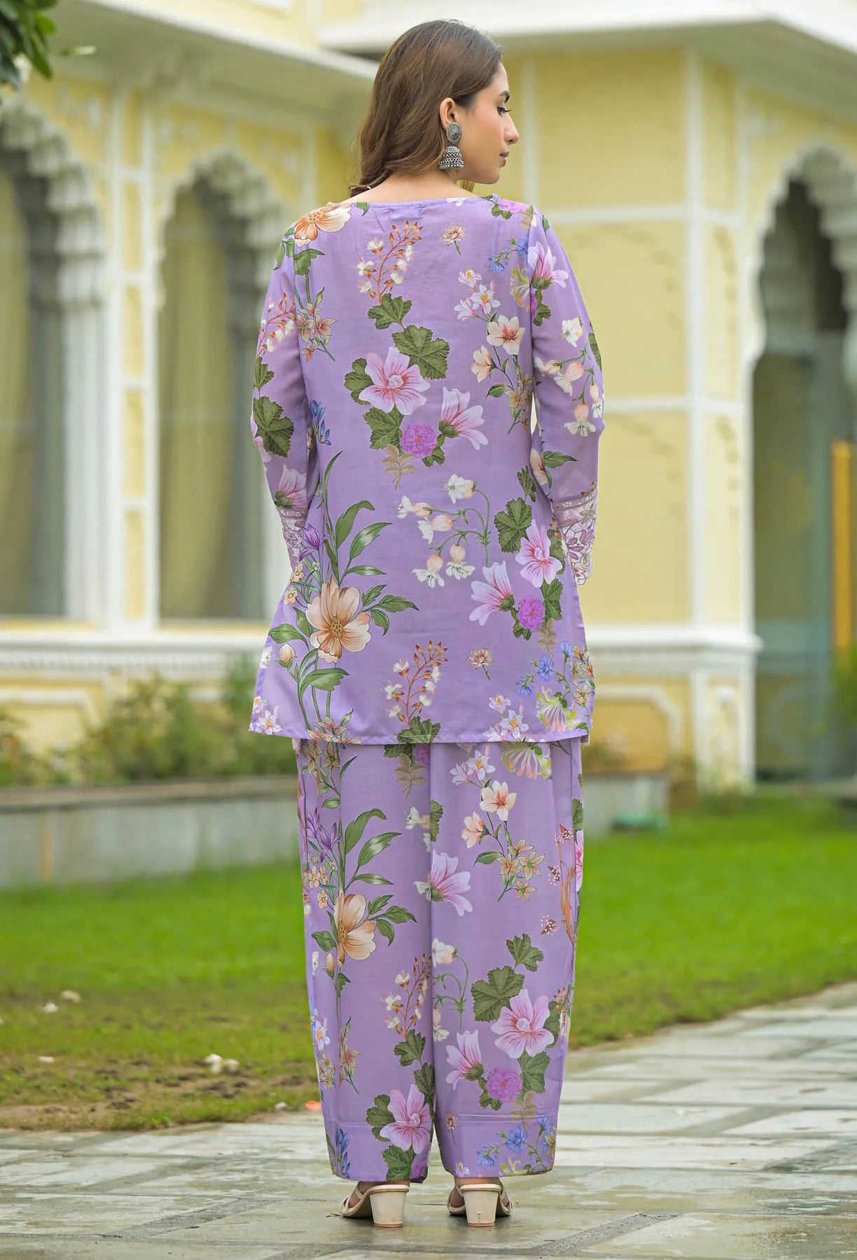 Woman wearing a purple floral outfit standing in front of a building with greenery.