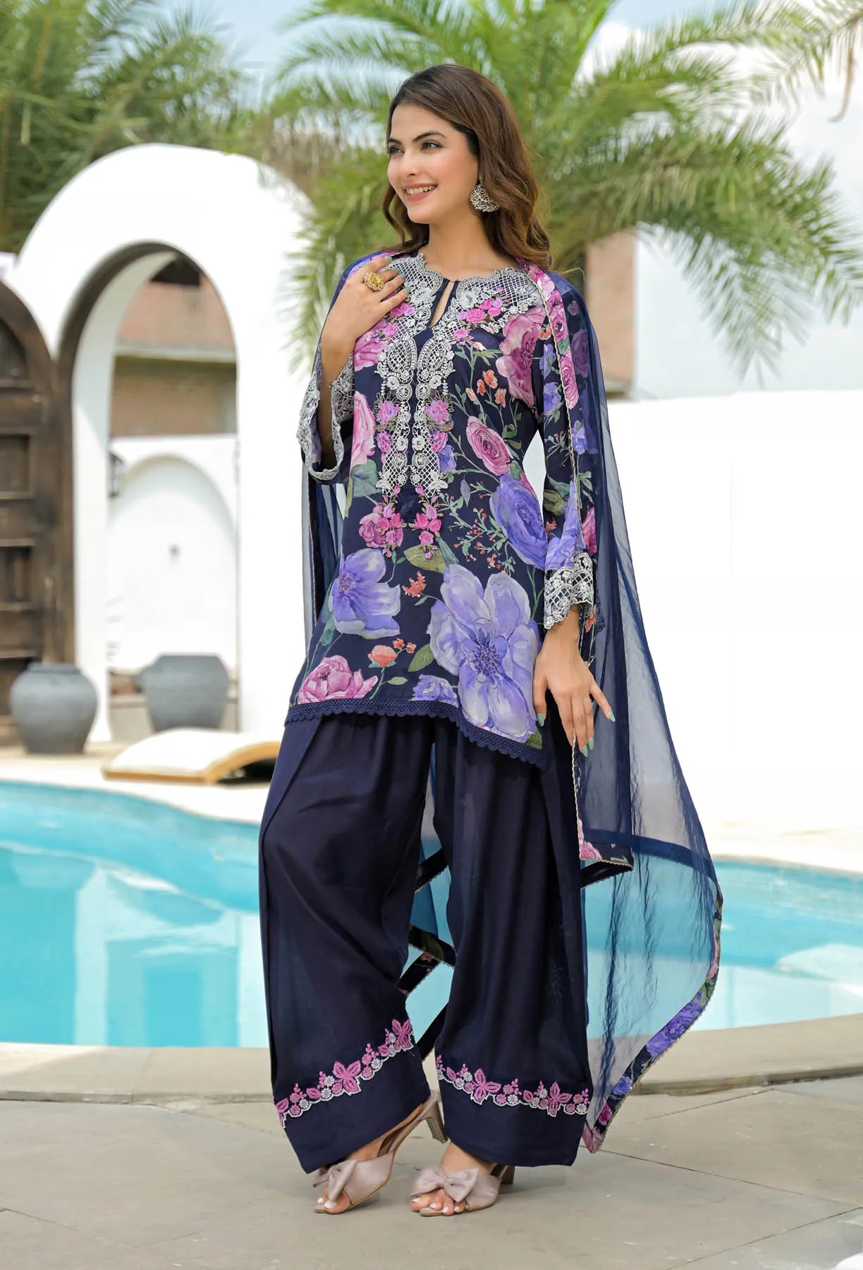 Woman in a floral embroidered outfit standing by a pool with palm trees in the background