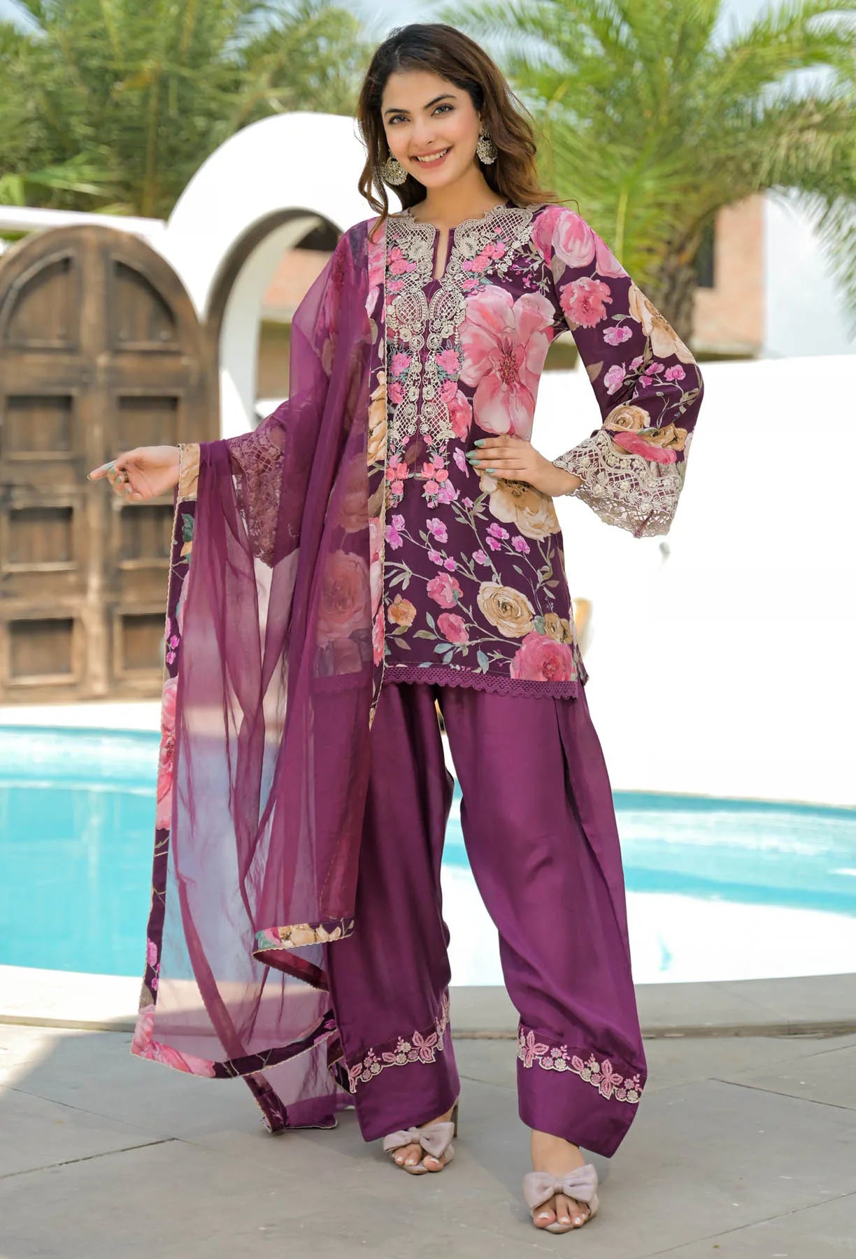 Woman in a floral purple outfit standing by a pool with palm trees in the background