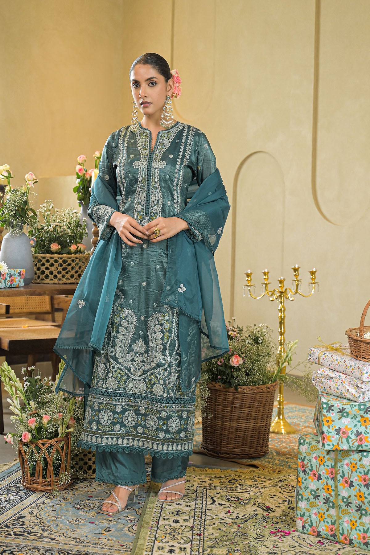 Woman in traditional teal embroidered outfit standing in a decorated room with flowers and decor.