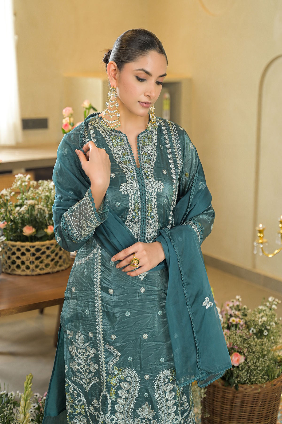 Woman wearing a teal embroidered outfit in an indoor setting with flowers and decor.