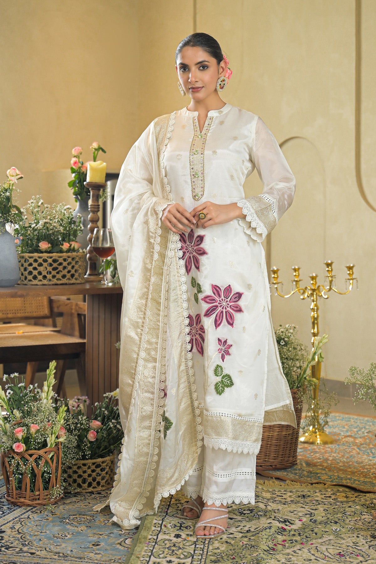 Woman in a white embroidered outfit standing in an elegant indoor setting with floral decorations.
