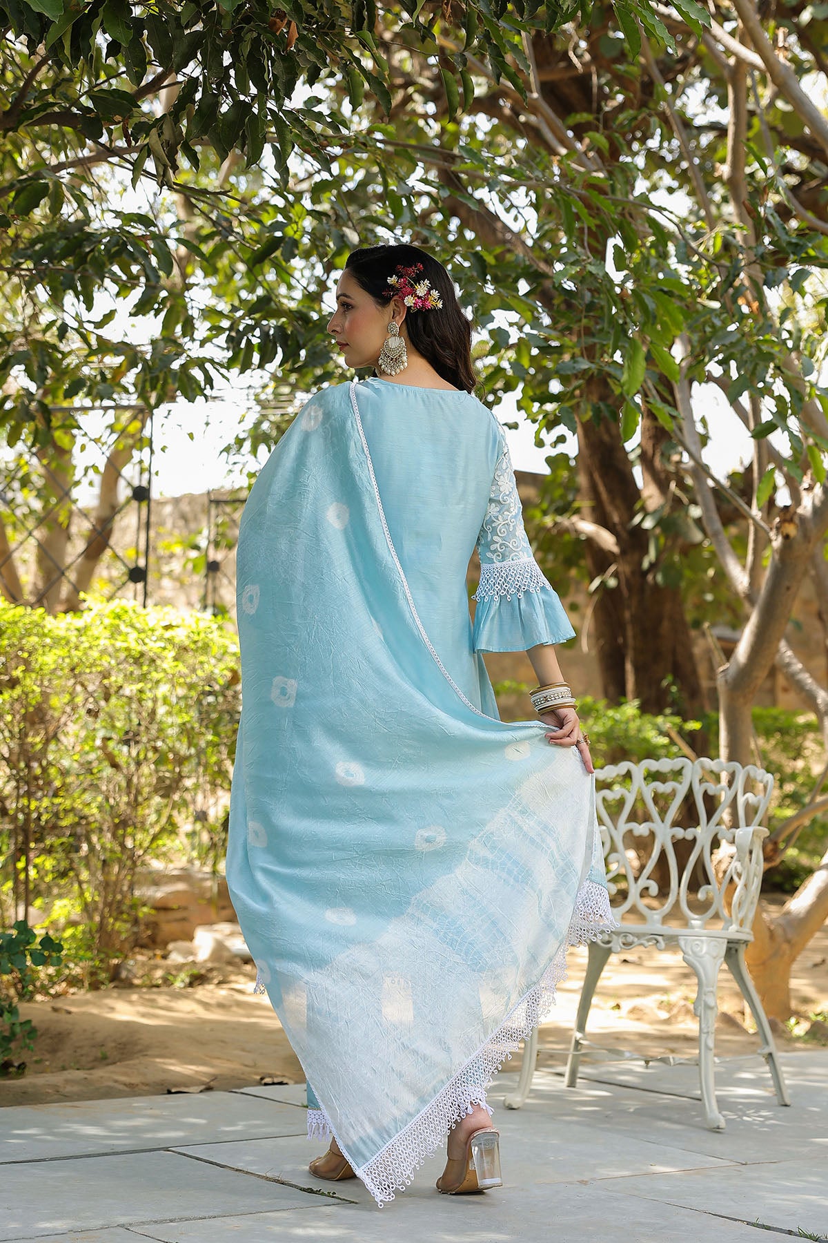 Woman in a light blue saree with white patterns walking outdoors near trees.