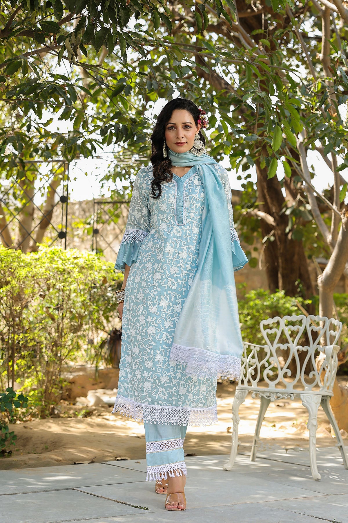 Woman in light blue traditional outfit standing outdoors with greenery and a bench in the background