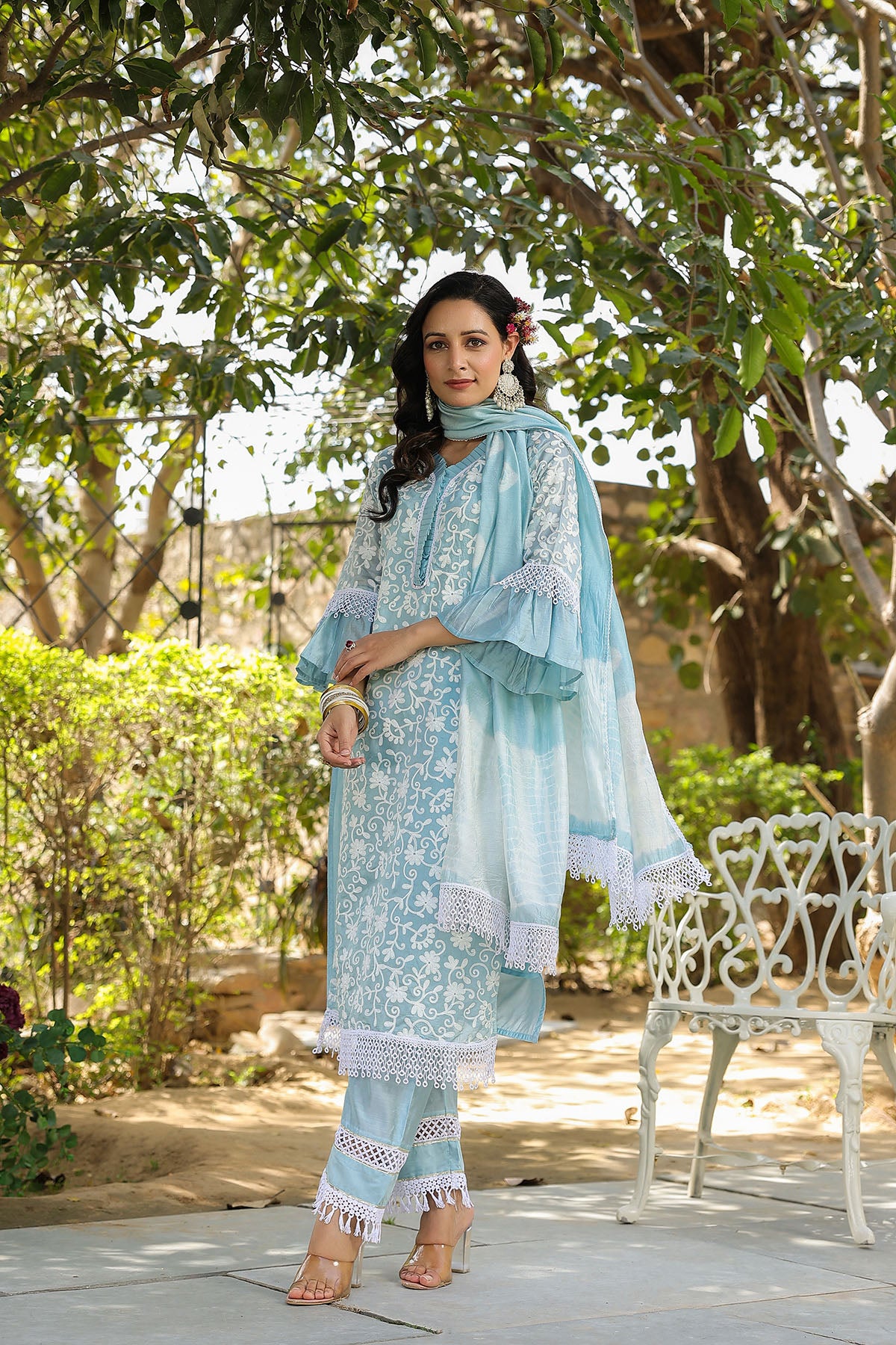 Woman in a light blue traditional outfit standing outdoors with greenery around