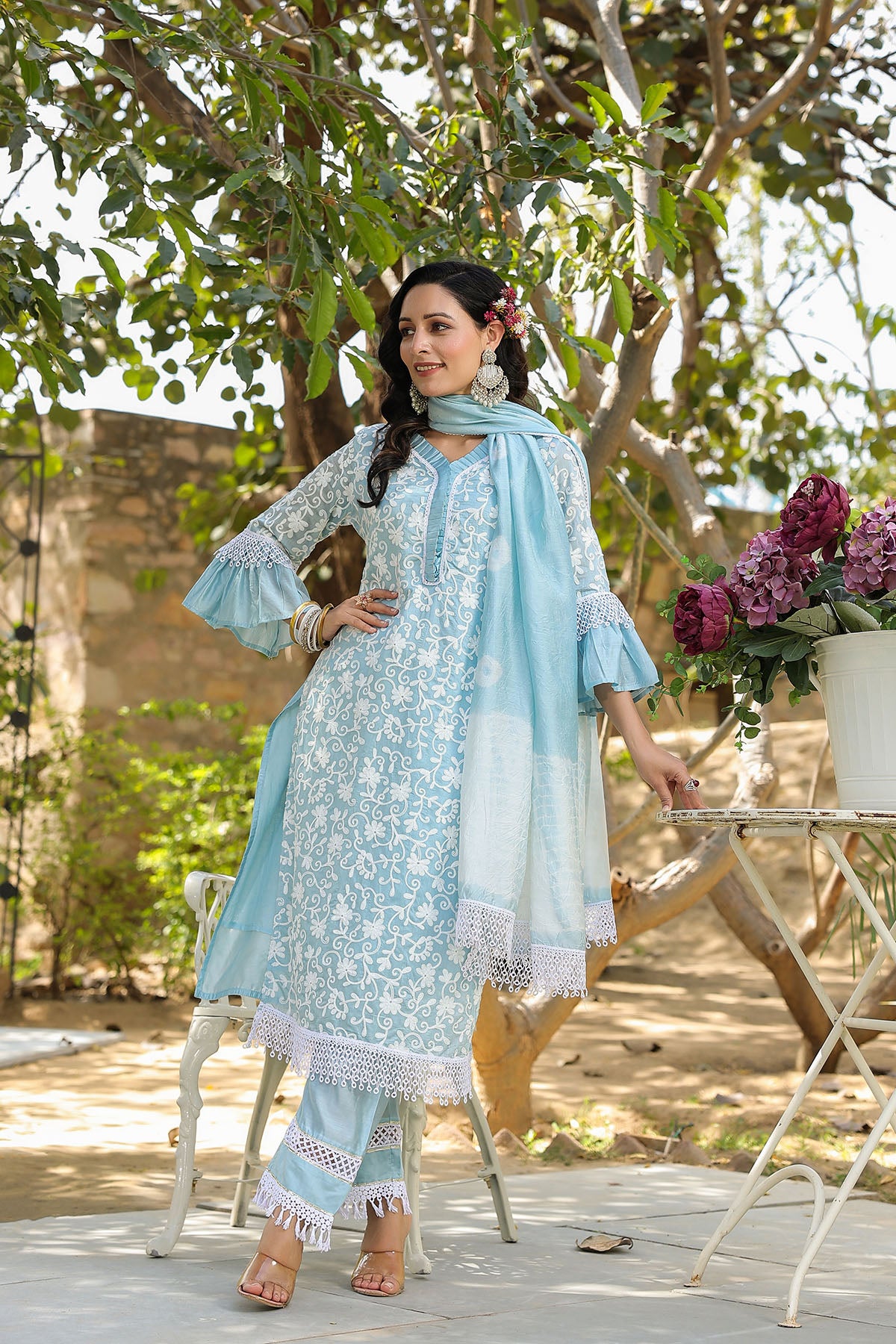 Woman in a light blue traditional outfit with a scarf, standing outdoors with flowers and a table.