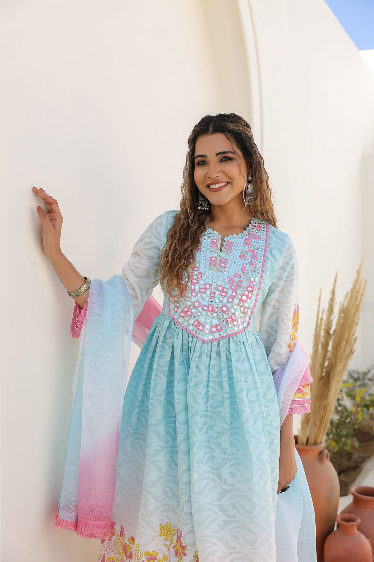 Woman wearing a light blue embroidered dress with a colorful dupatta, standing against a white wall with decorative pots.