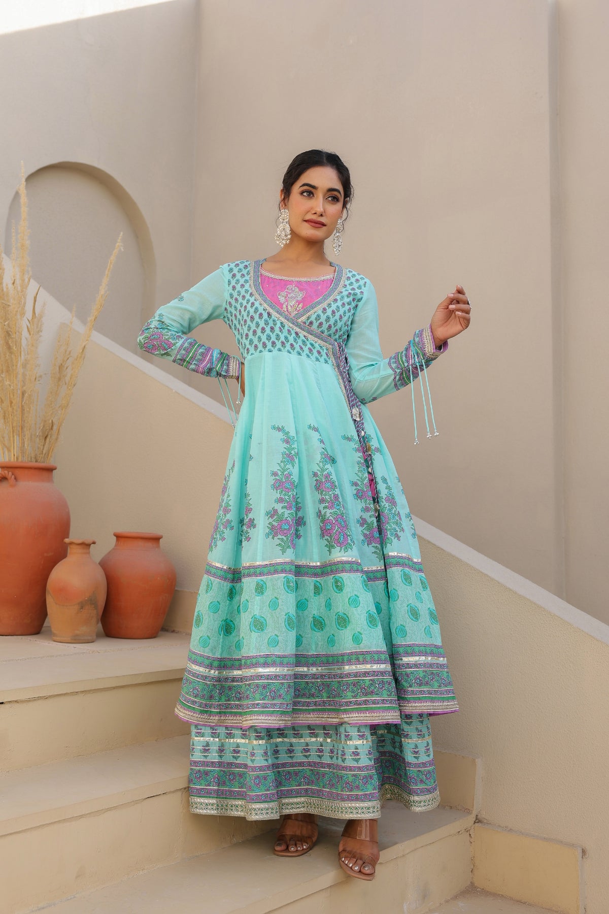 Woman in a light blue traditional outfit standing on steps with decorative pots in the background.
