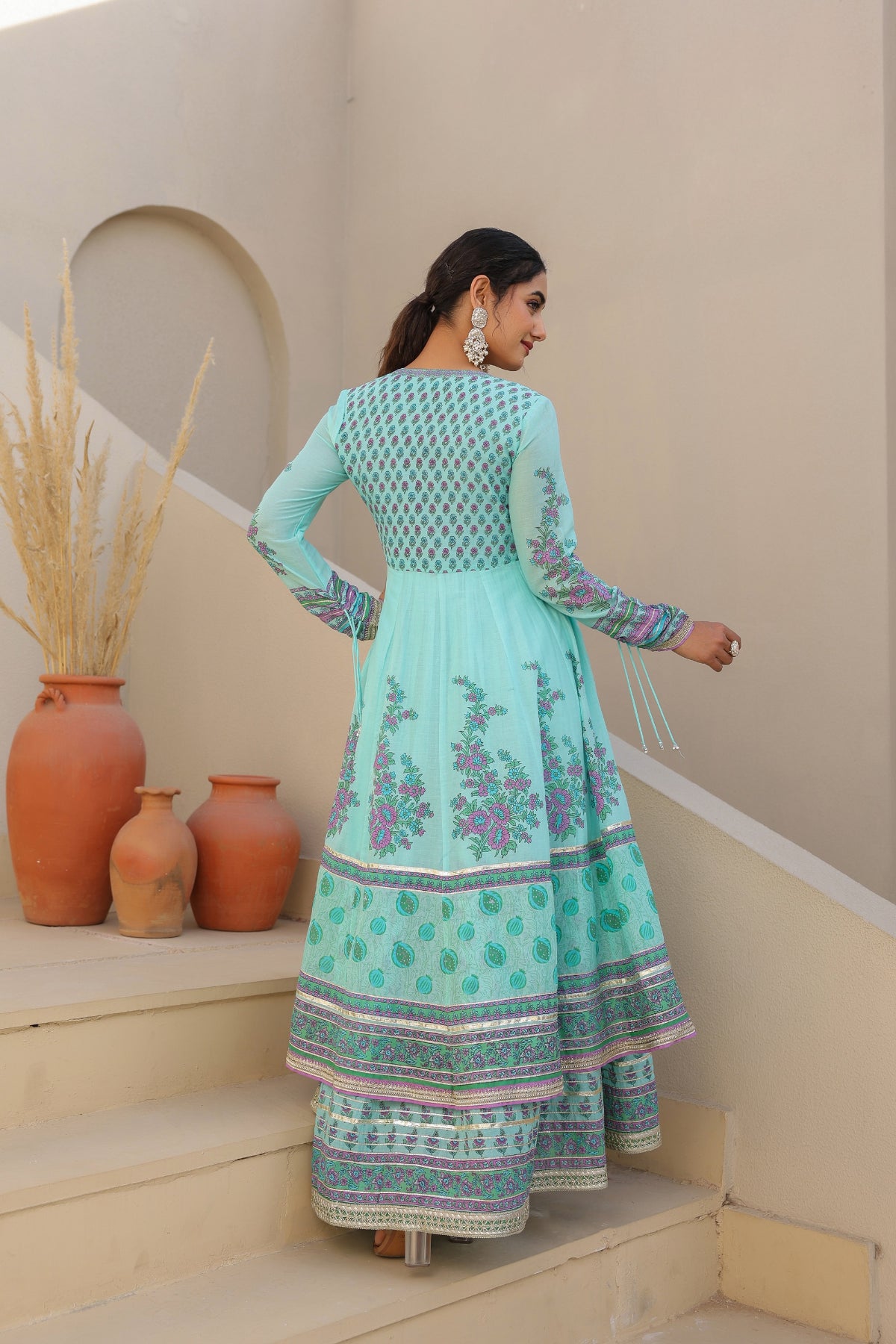 Woman in a turquoise traditional outfit standing on steps with decorative vases in the background.
