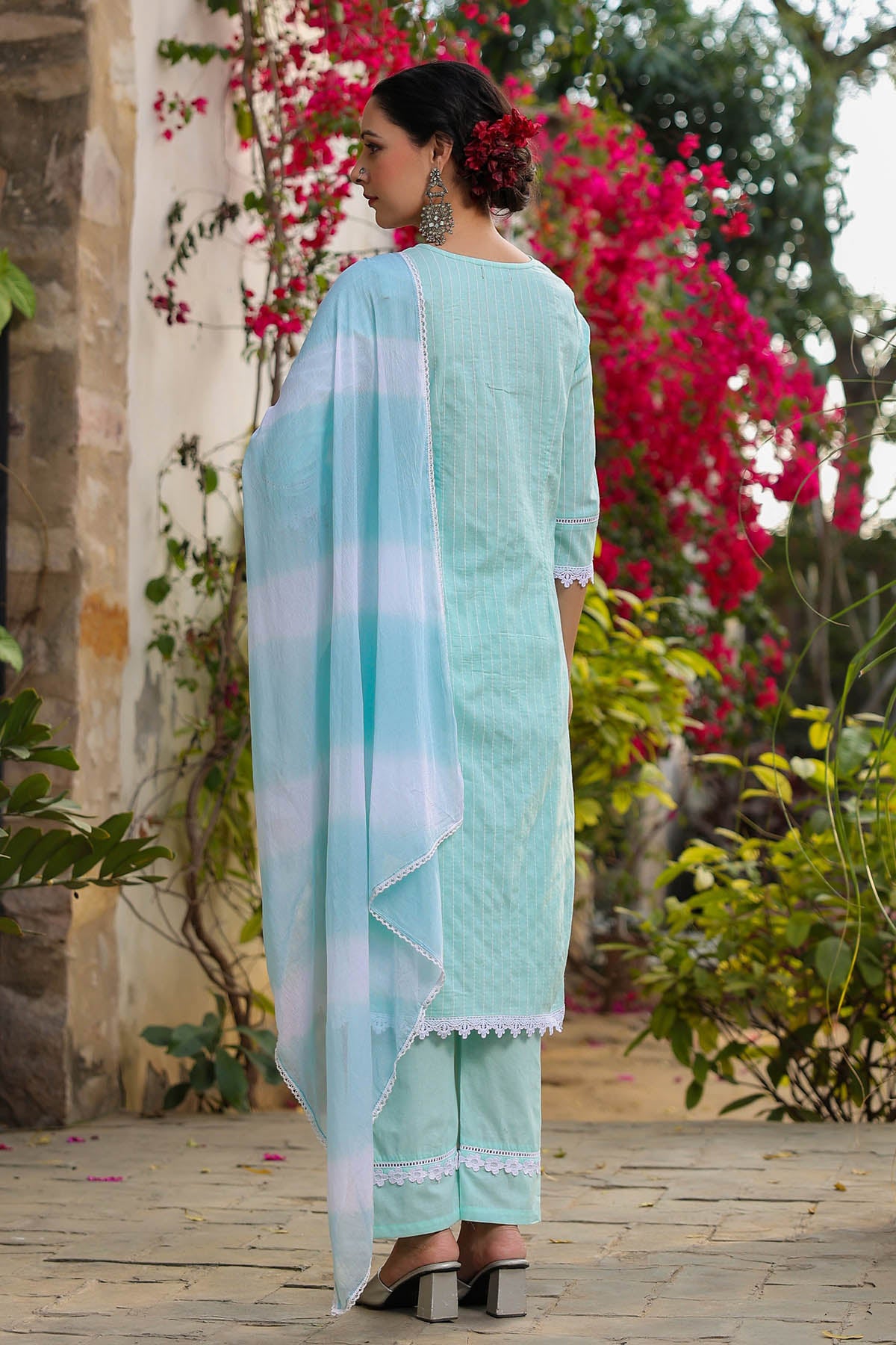 Woman in a light blue traditional outfit standing in a garden with pink flowers and greenery.