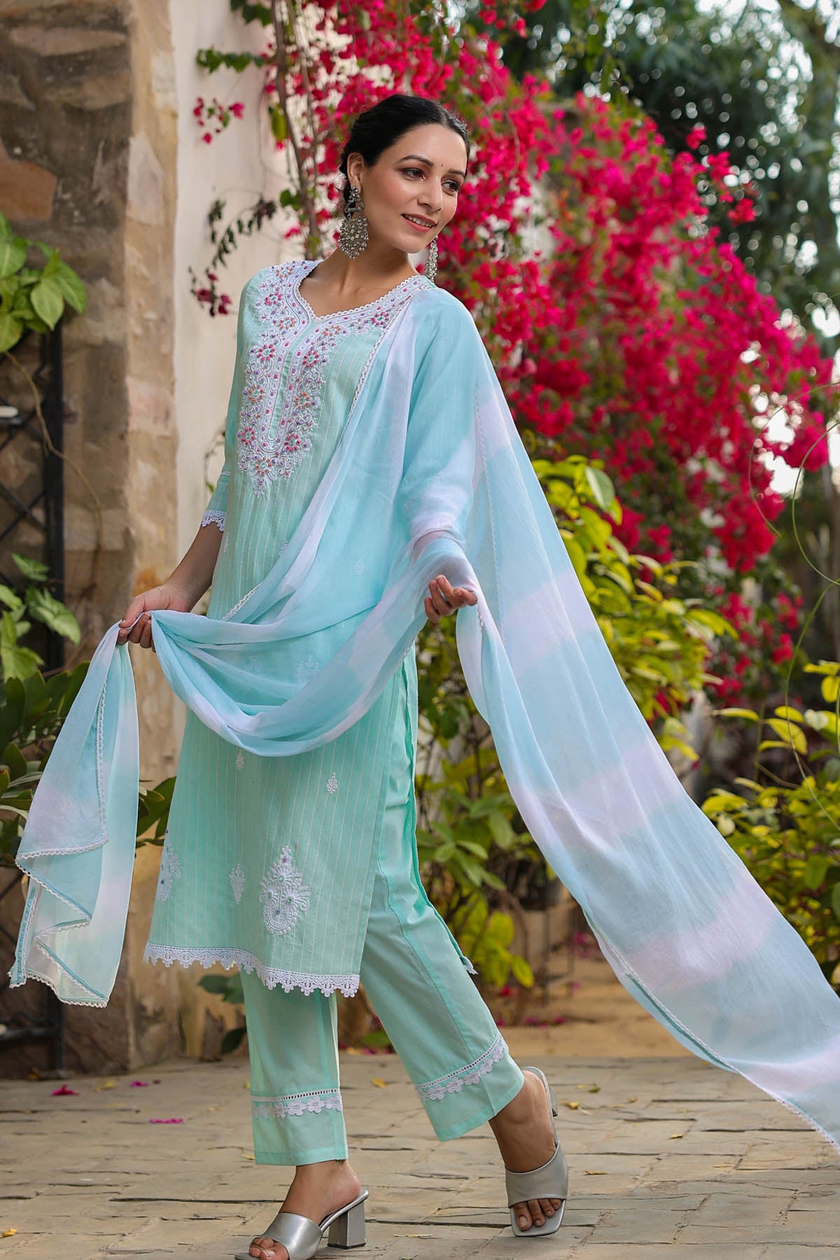Woman in a light blue traditional outfit with a matching dupatta, standing outdoors with flowers in the background.