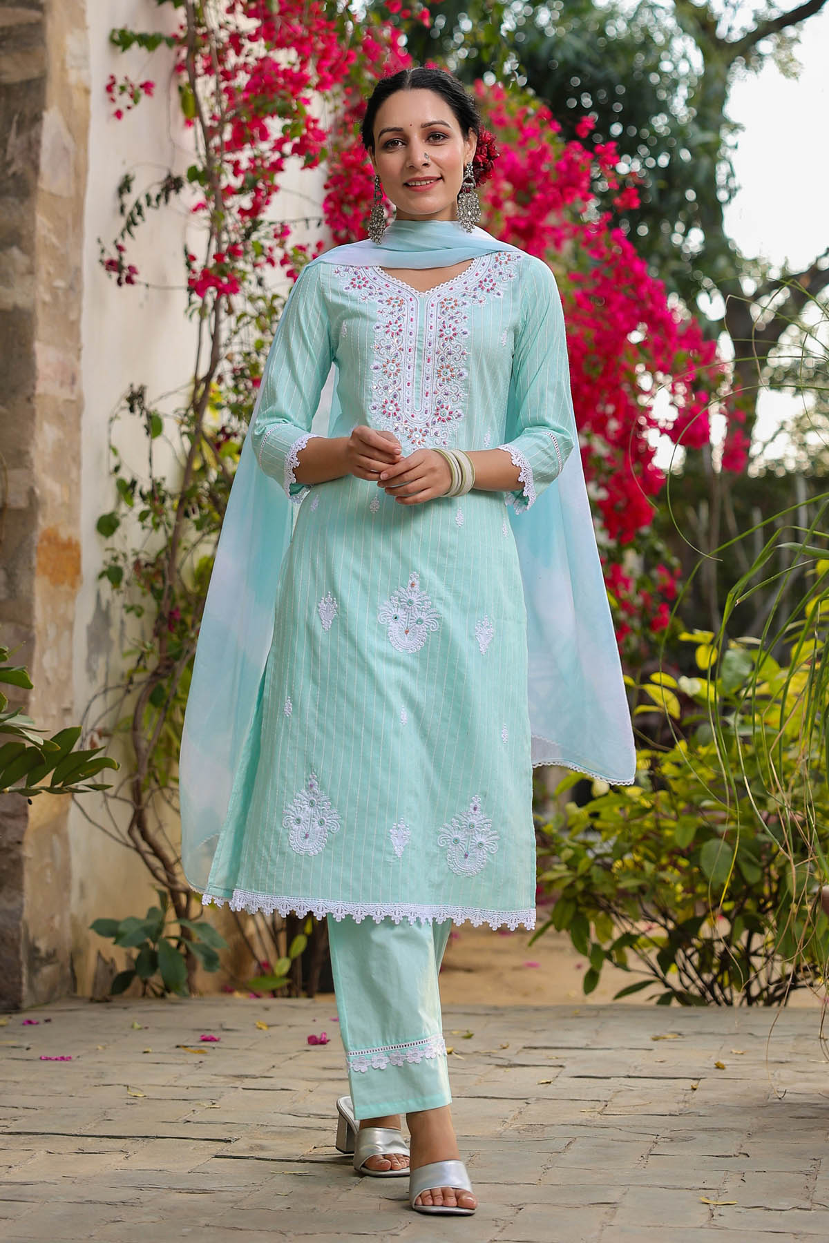 Woman in a light blue traditional outfit standing outdoors with flowers in the background
