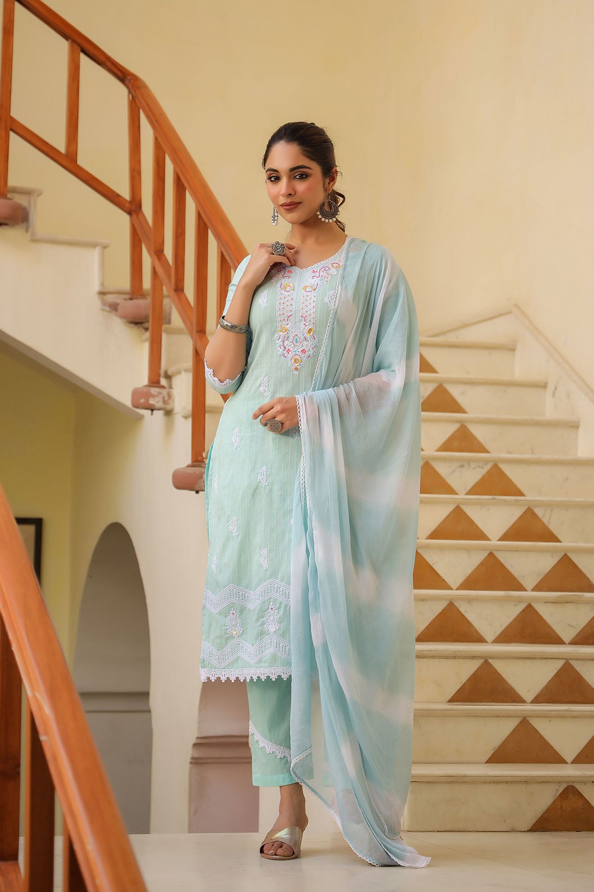 Woman in a light blue traditional outfit standing in a home setting with a staircase.