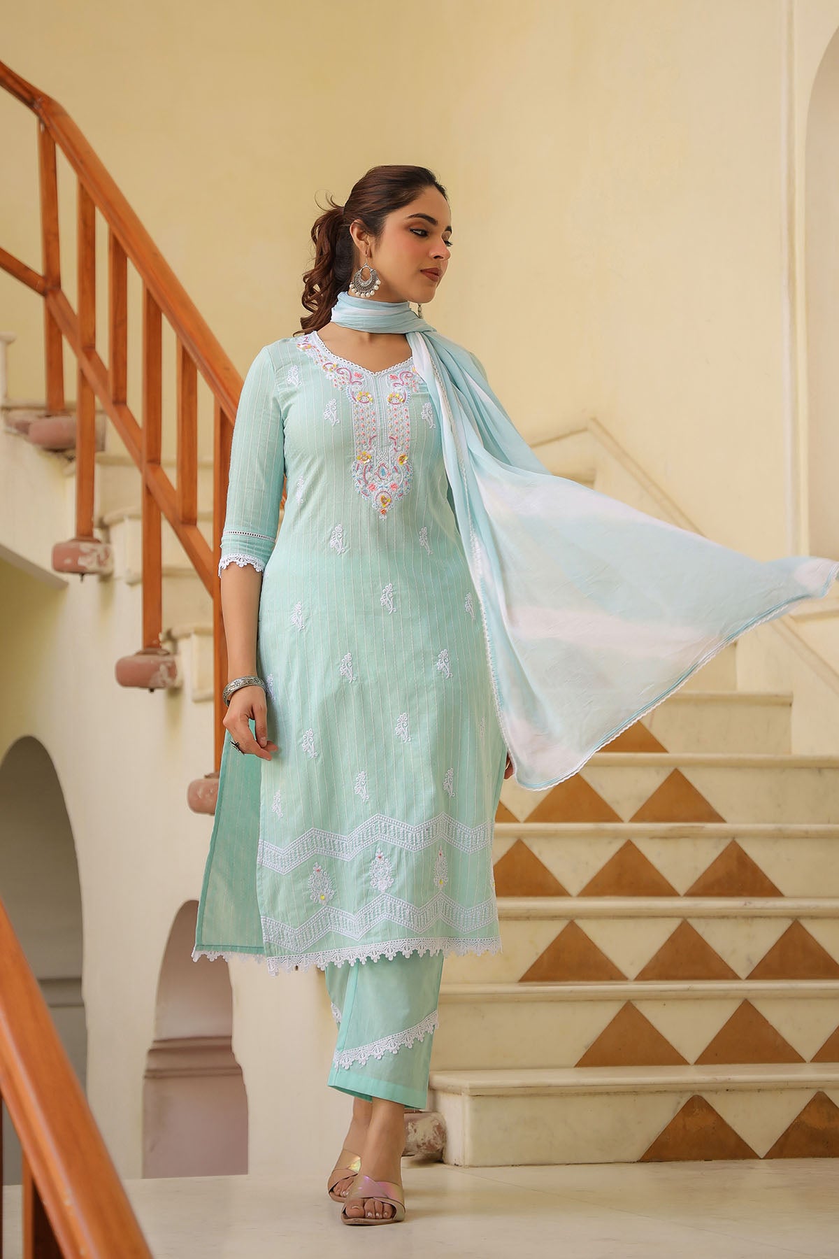 Woman in a light green traditional outfit with a white dupatta standing in front of a staircase.