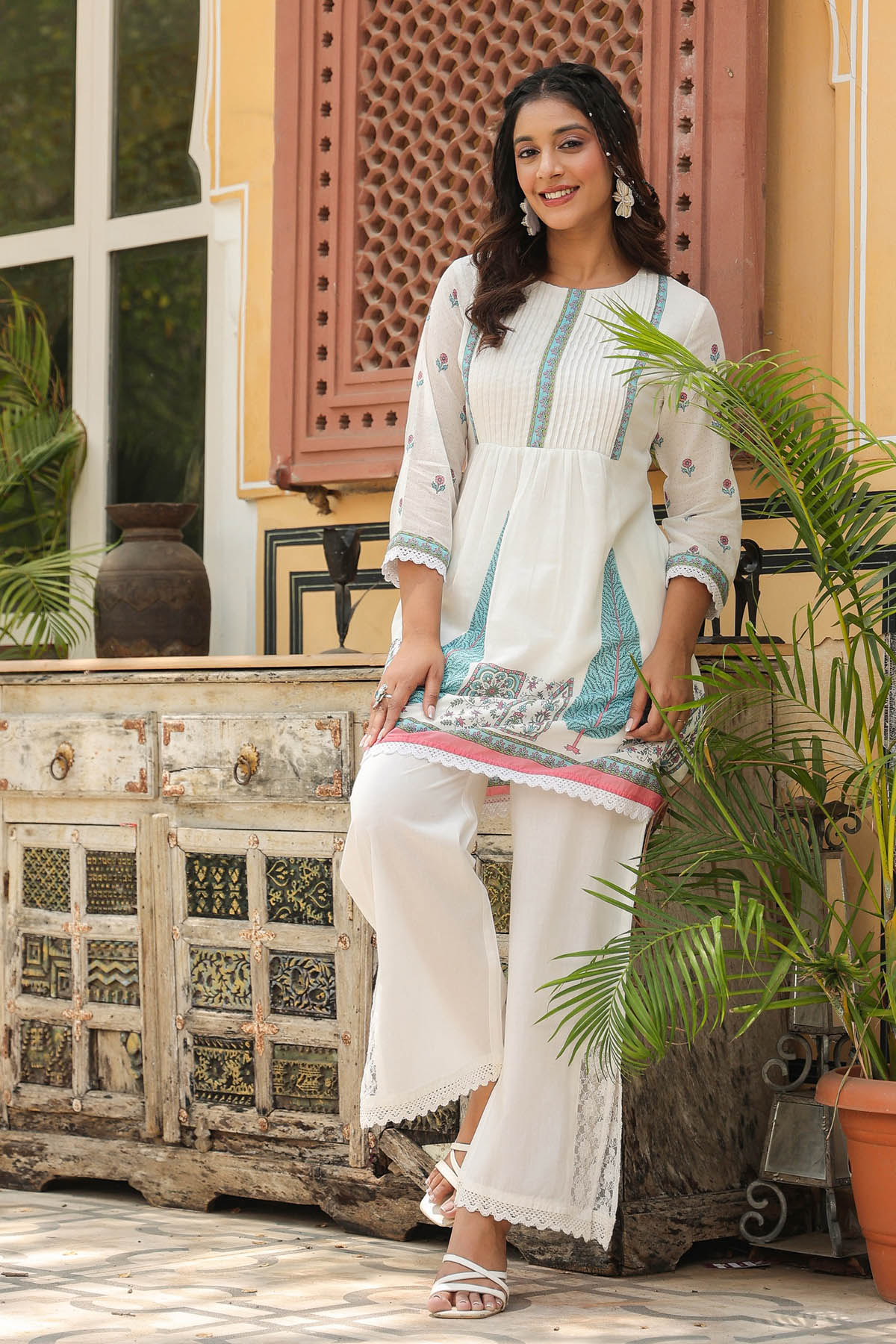 Woman in a traditional outfit standing in front of a decorative wall with plants.