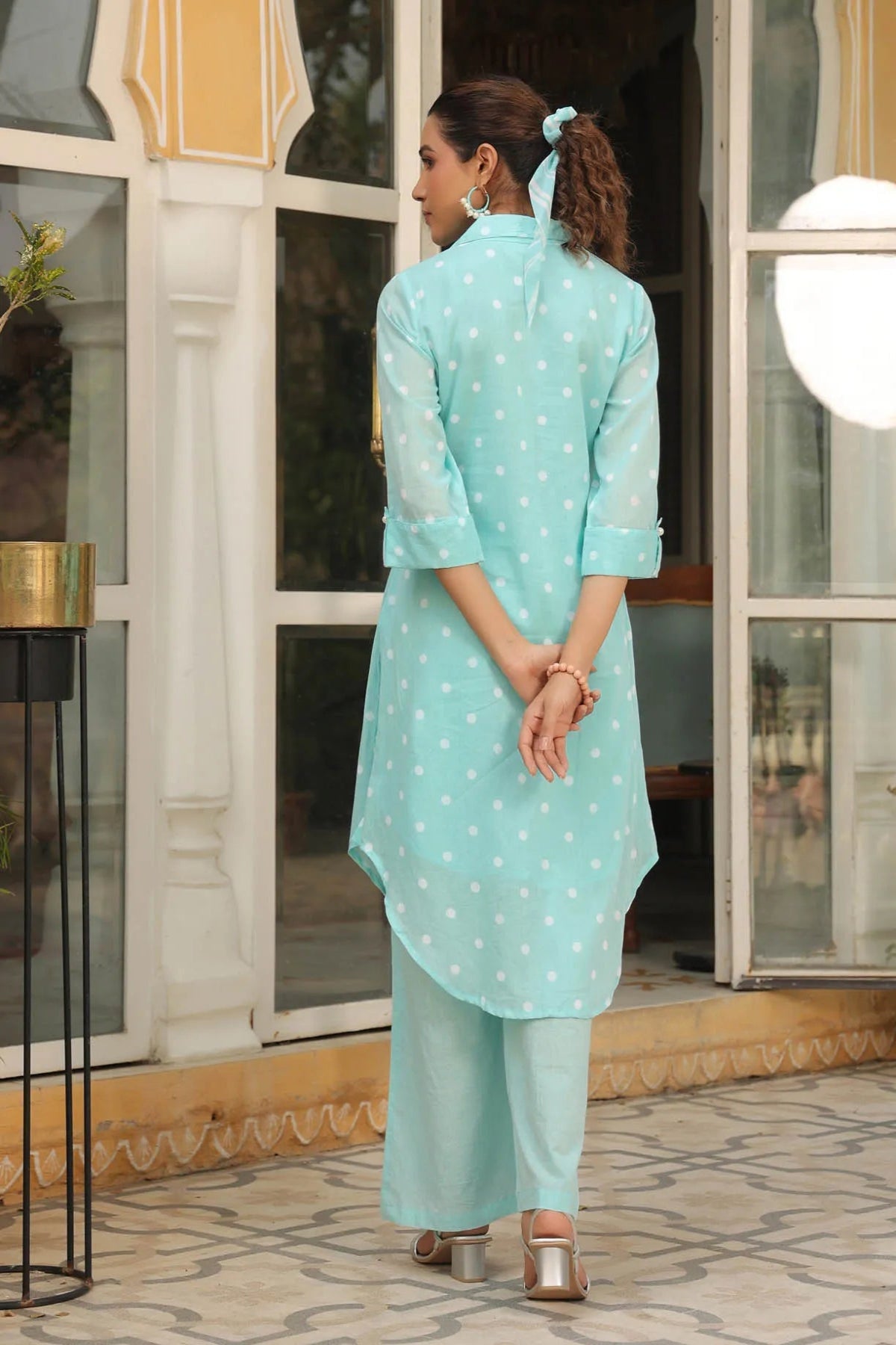 Woman wearing a light blue traditional outfit with white patterns, standing in an outdoor setting.
