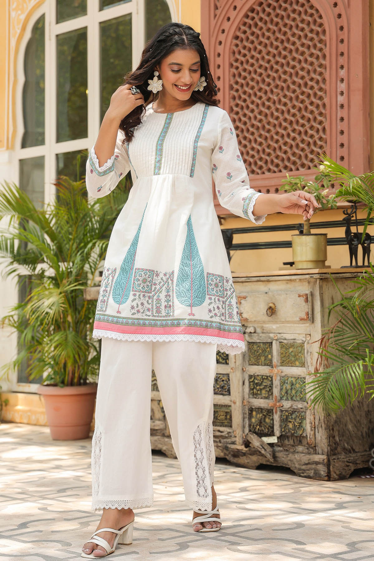 Woman in a white and blue embroidered outfit standing outdoors with plants and a decorative wall in the background.