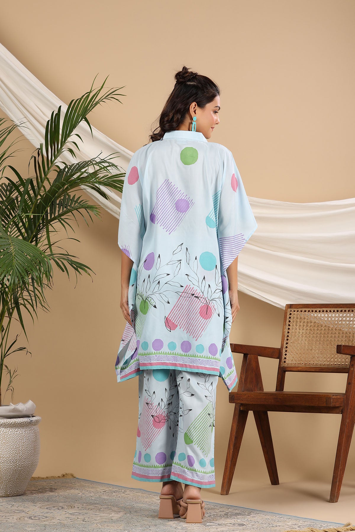 Woman wearing a colorful outfit with a patterned shirt and pants in a room with a plant and wooden chair.