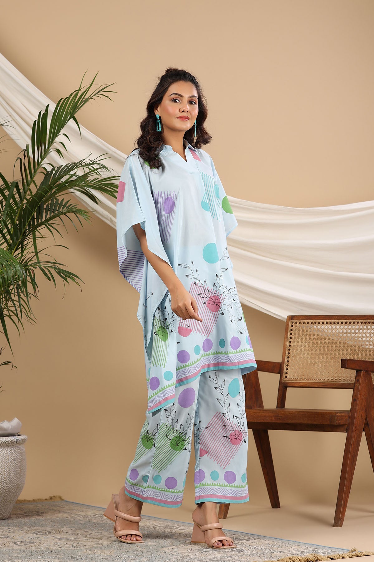 Woman wearing a light blue outfit with colorful patterns in a room with a plant and wooden chair.