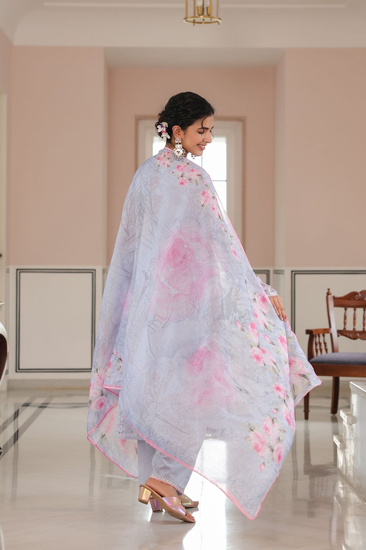 Woman wearing a light blue floral dress in a room with pink walls and a wooden chair.