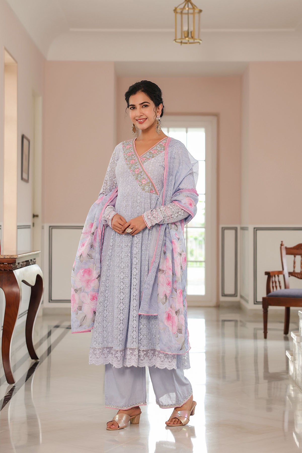 Woman in a light gray traditional outfit with pink floral patterns standing in a well-lit room.