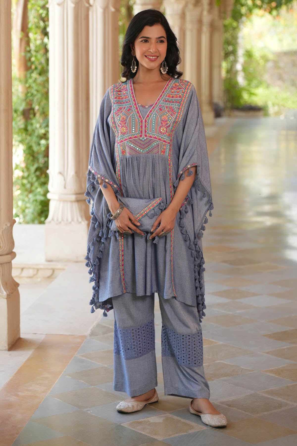Woman wearing a gray embroidered outfit standing in a sunlit outdoor setting with columns.