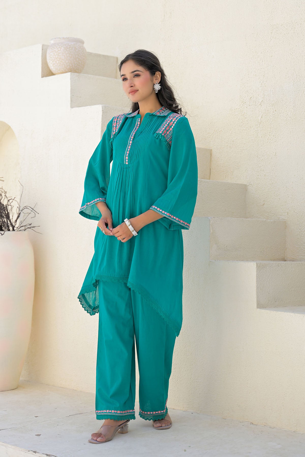 Woman wearing a teal traditional outfit with white embroidery in a minimalistic setting.