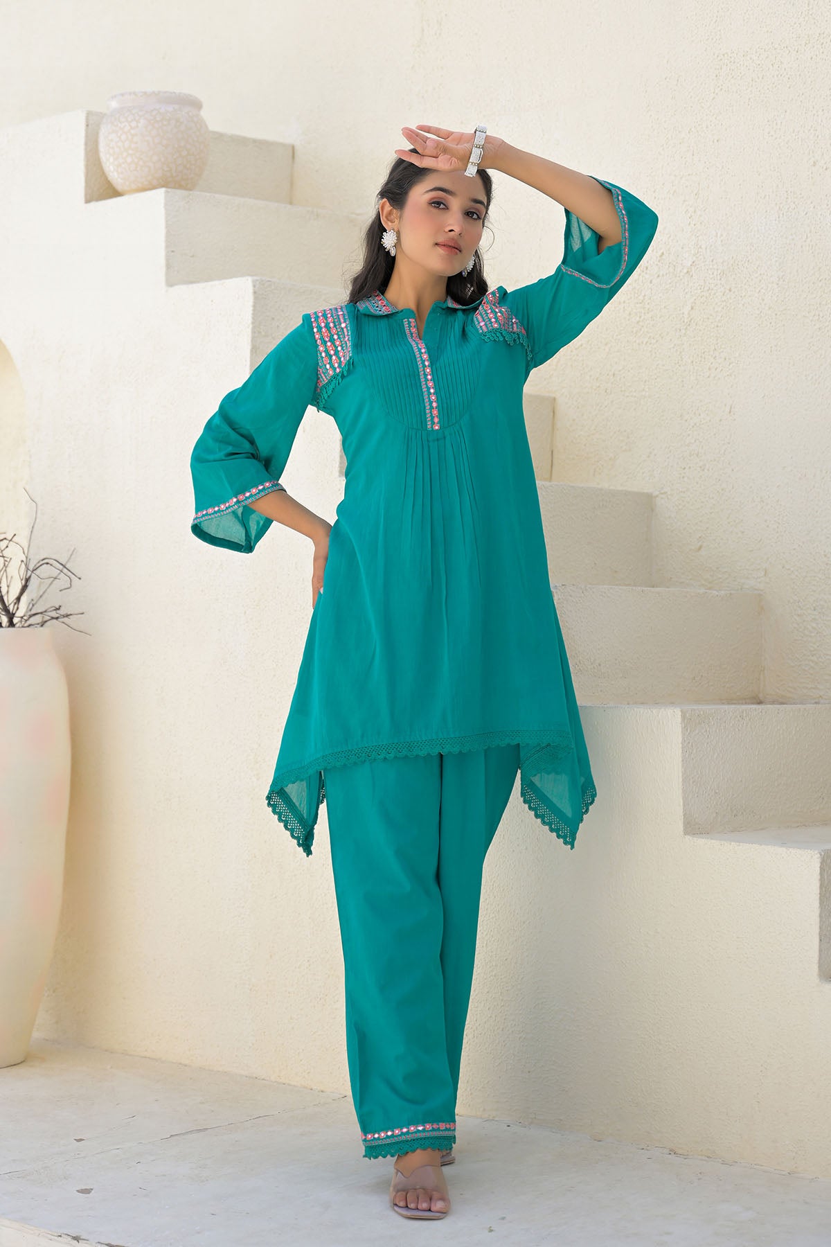 Woman wearing a teal traditional outfit with white embroidery in a minimalistic setting.