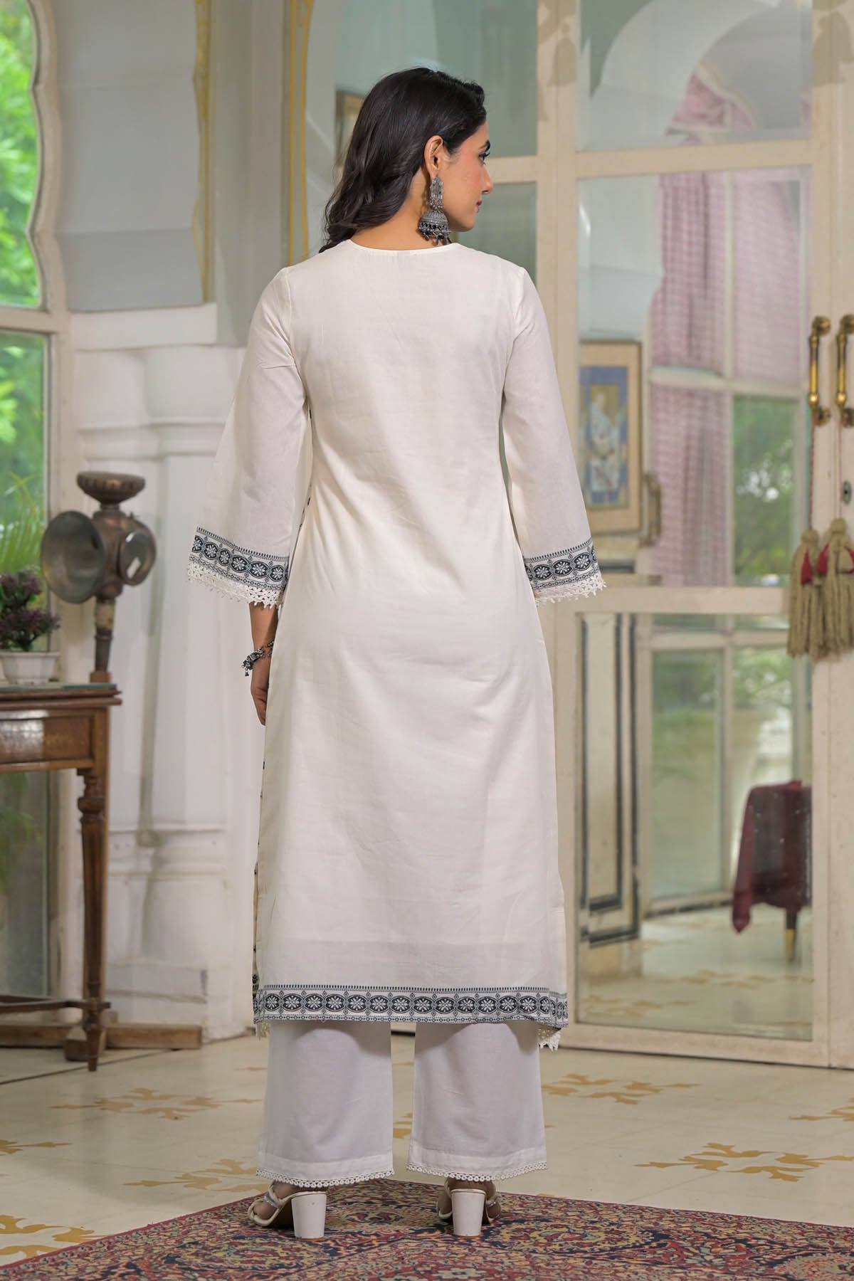 Woman wearing a white traditional outfit with blue embroidery in a decorated room.