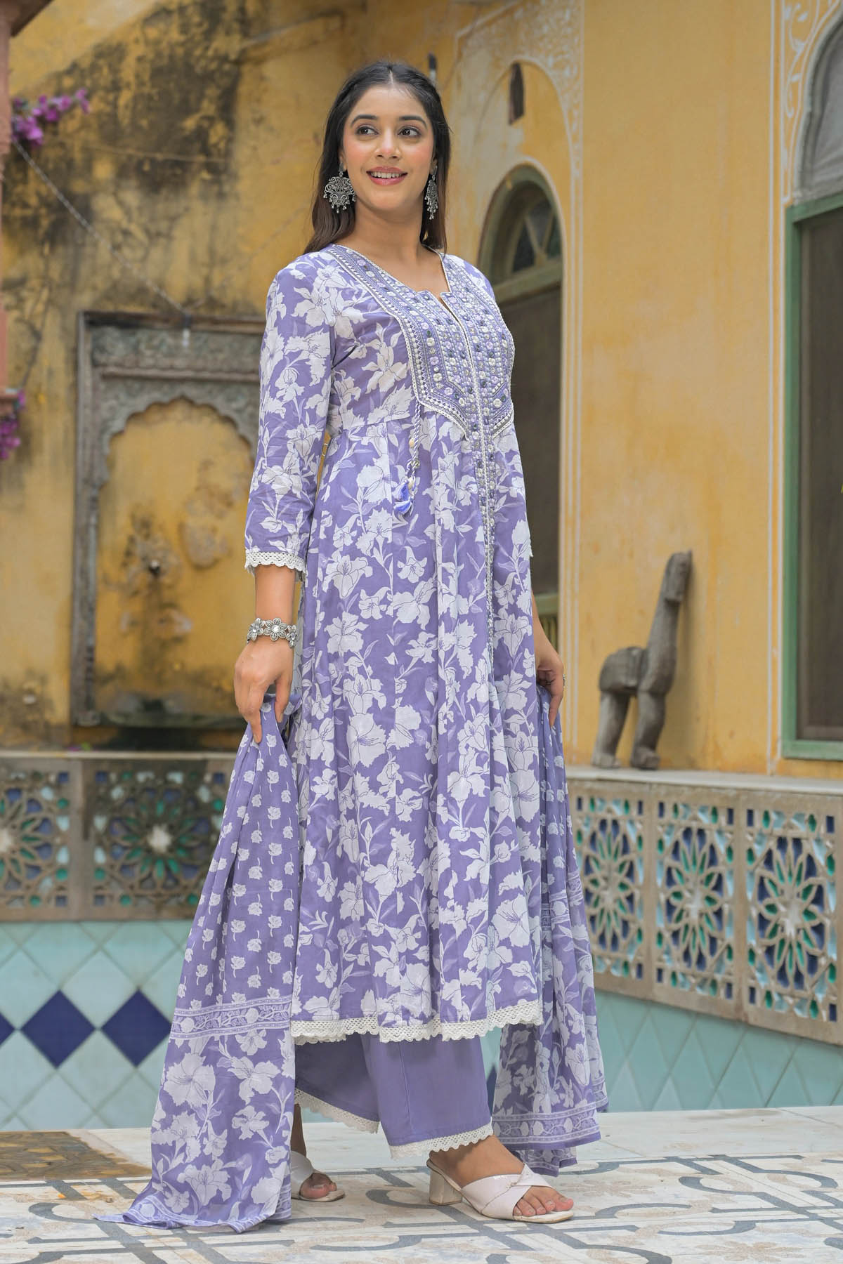 Woman wearing a purple floral outfit standing in front of a decorative wall.