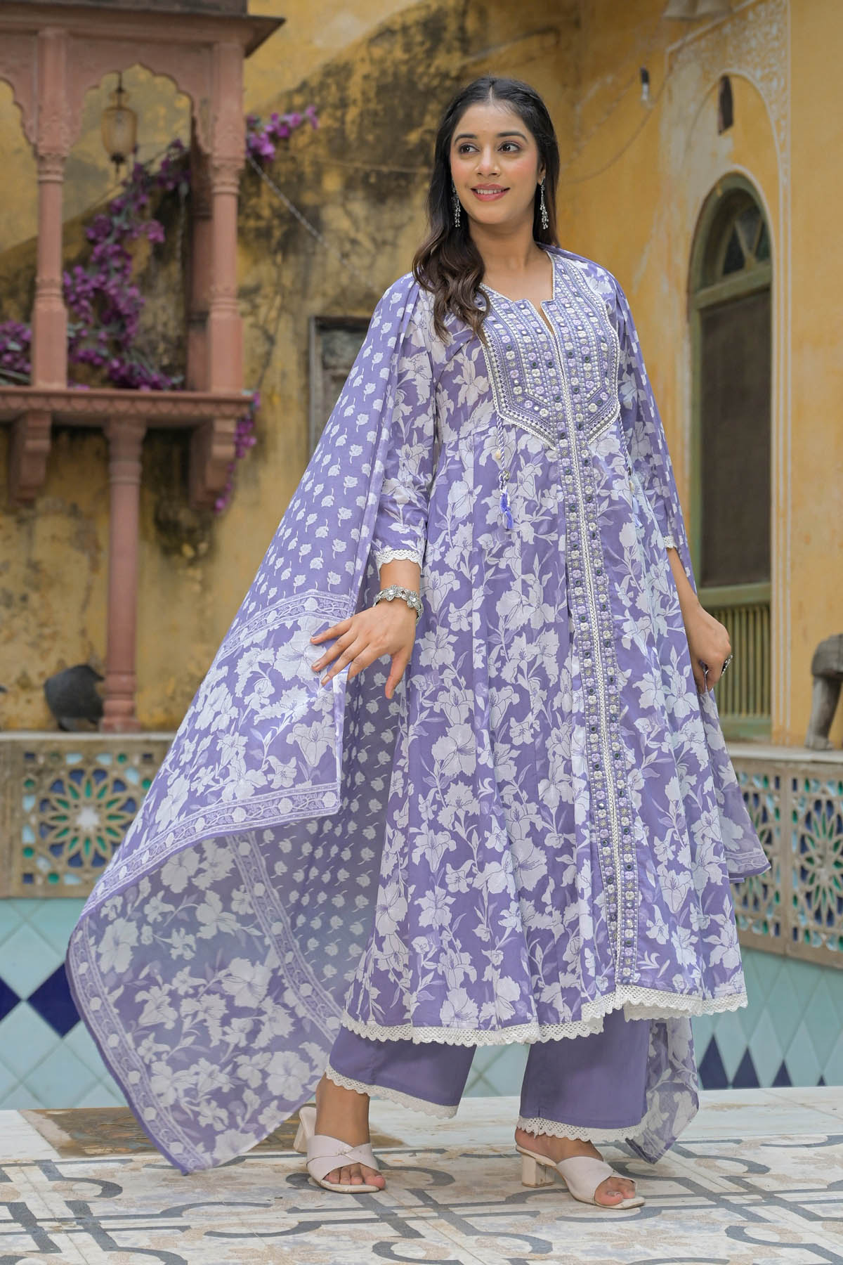 Woman wearing a purple floral traditional outfit in an outdoor setting with decorative architecture.