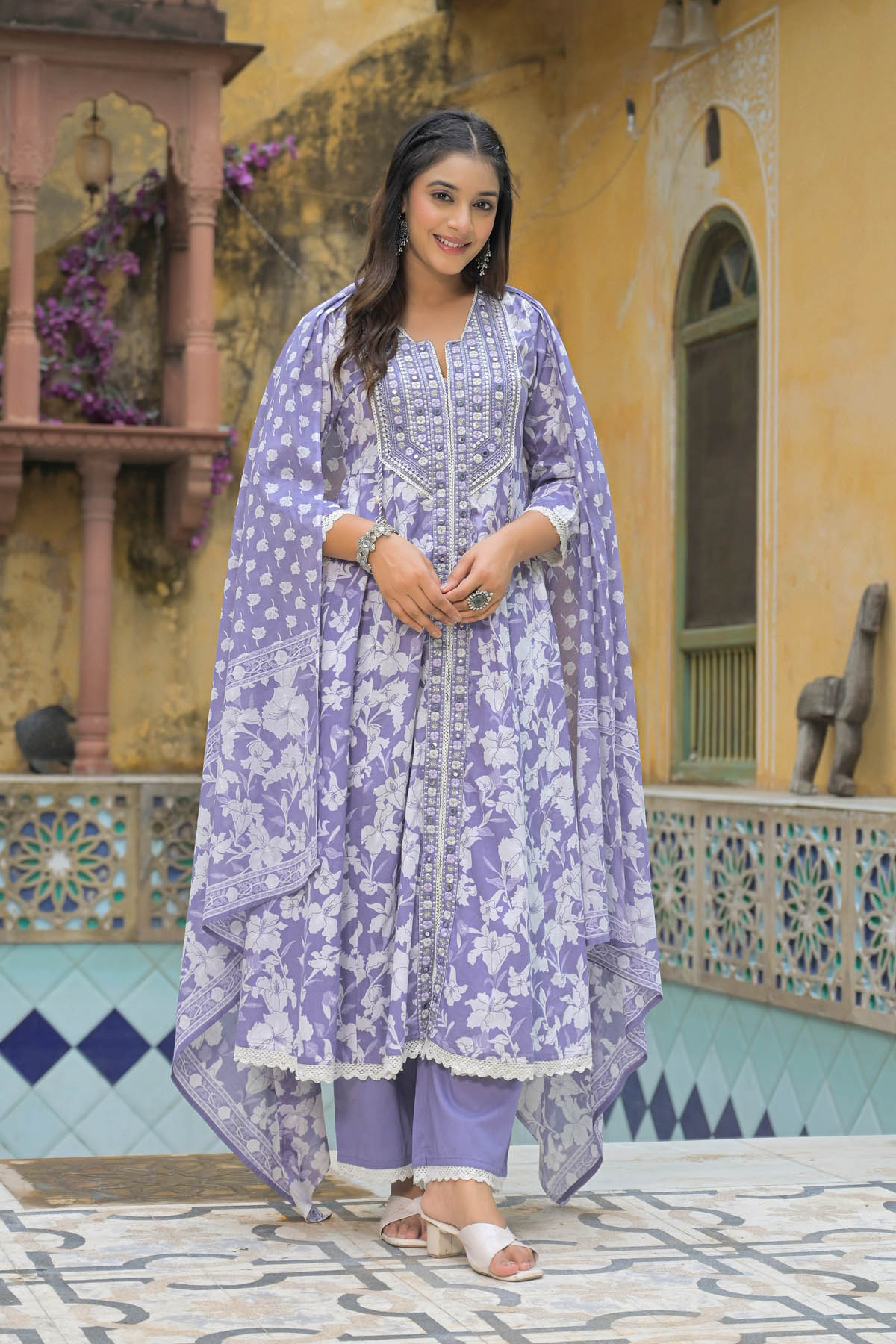 Woman in a purple floral traditional outfit standing in front of a colorful architectural background.