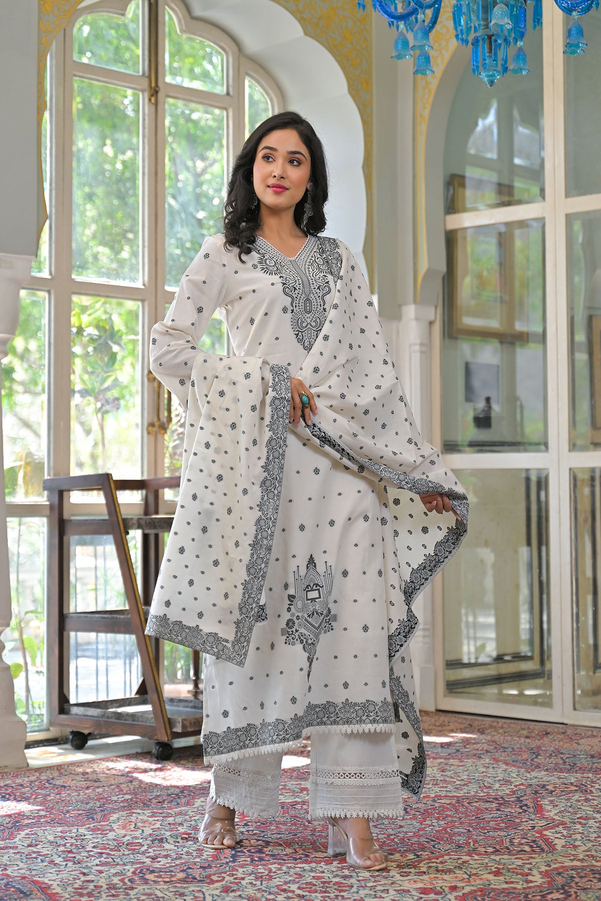 Woman in a white and gray traditional outfit standing in a room with large windows and a chandelier.