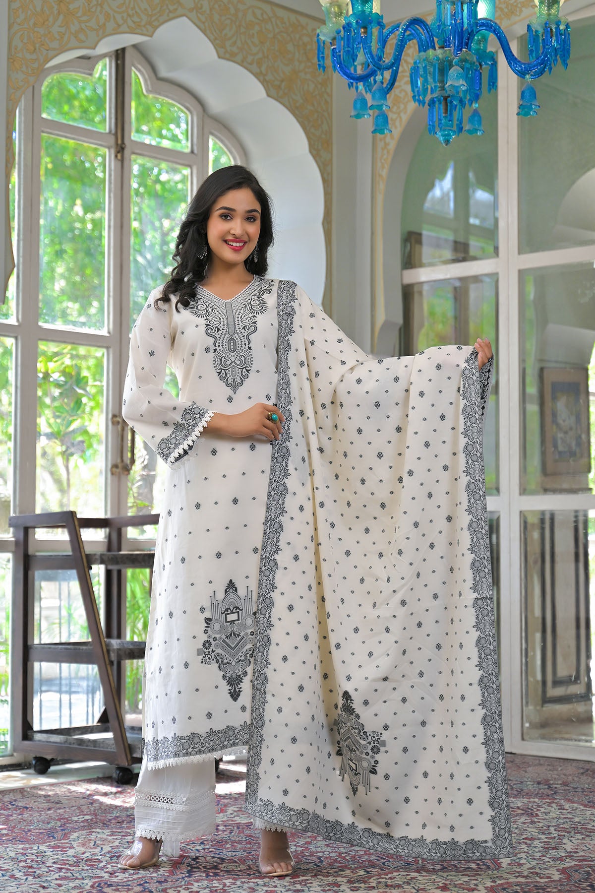 Woman in a white and gray embroidered traditional outfit standing in a room with large windows.