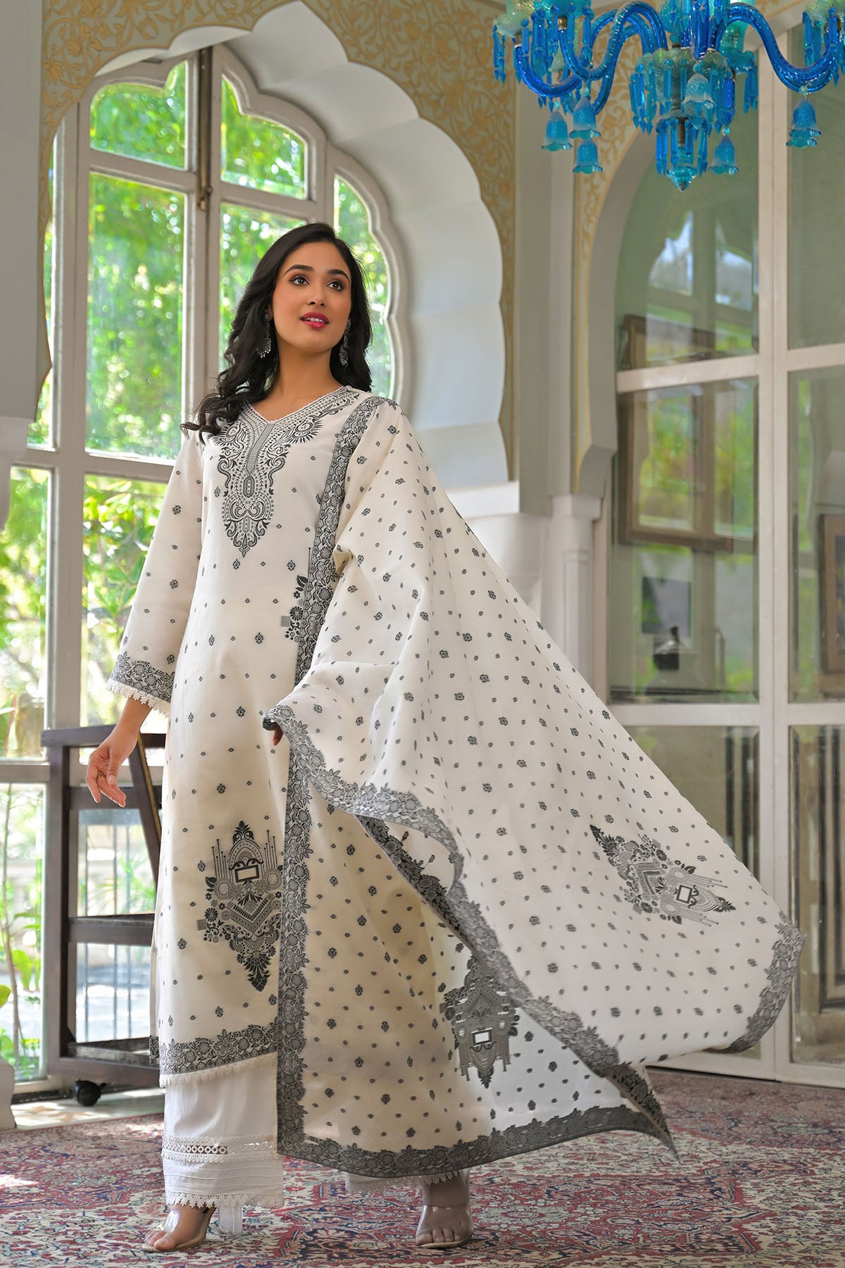Woman in a white and gray traditional outfit standing in a room with large windows and a blue chandelier.