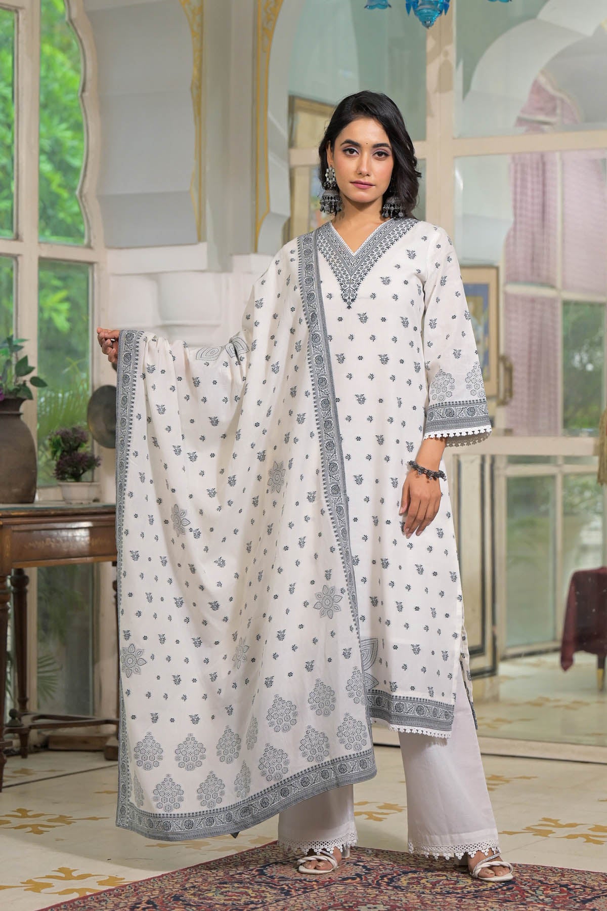 Woman in a white traditional outfit with intricate patterns standing in a decorated room.