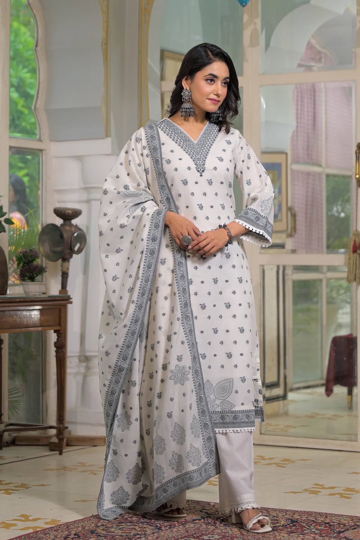 Woman in a white traditional outfit with blue patterns standing in an elegant room.