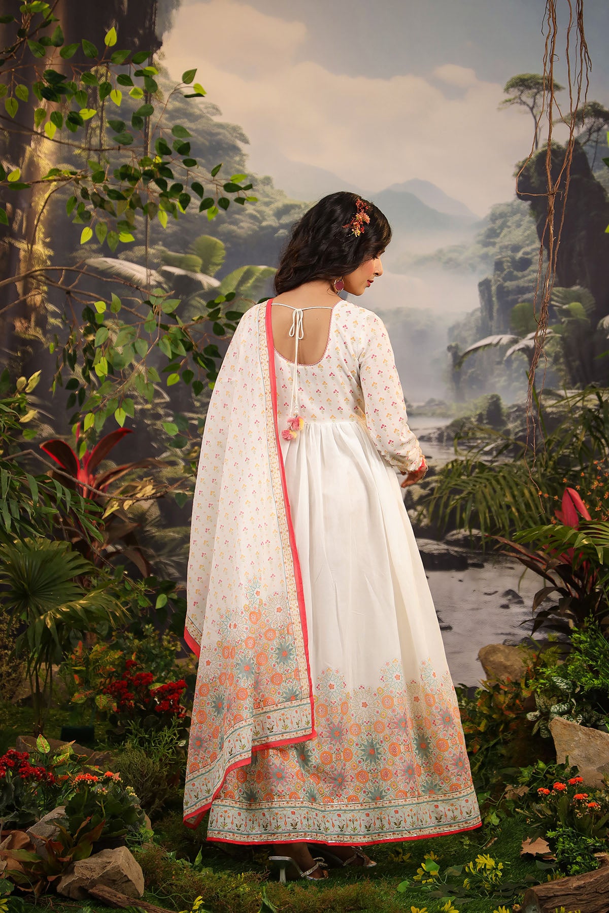 Woman in a white traditional outfit with floral patterns standing in a lush, tropical setting with a waterfall.