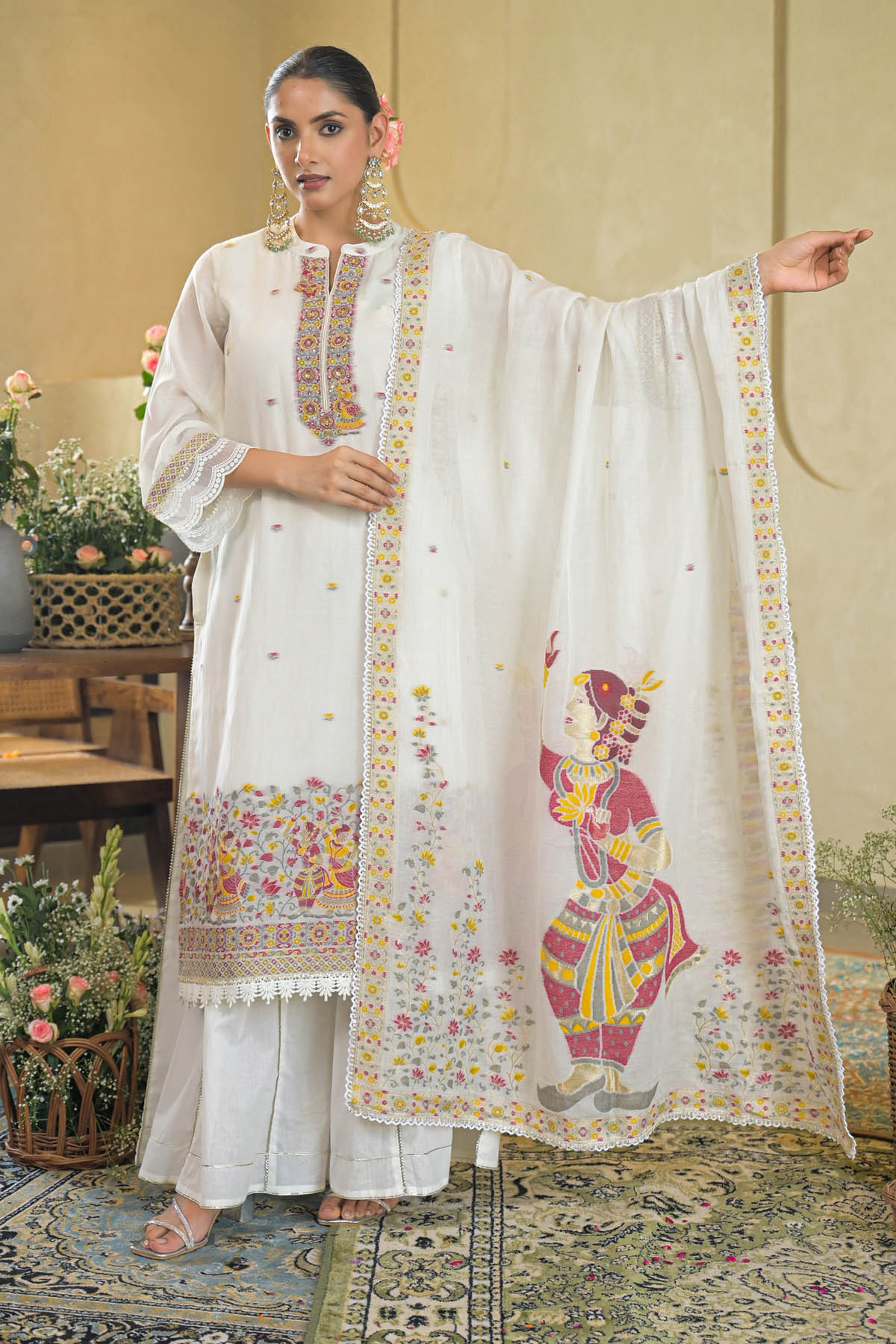 Woman wearing a traditional white embroidered outfit with a colorful design, standing in a decorated room.