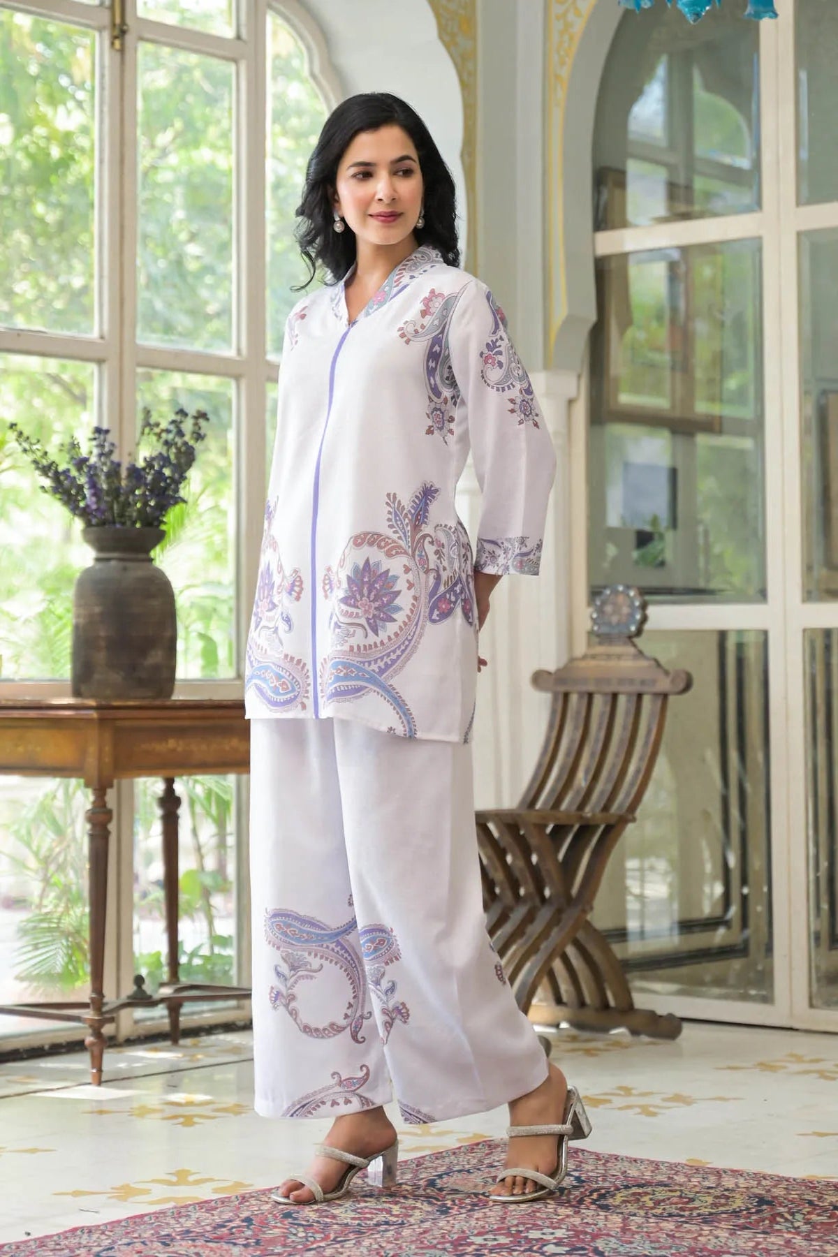 Woman wearing a white outfit with purple floral patterns in a room with large windows.